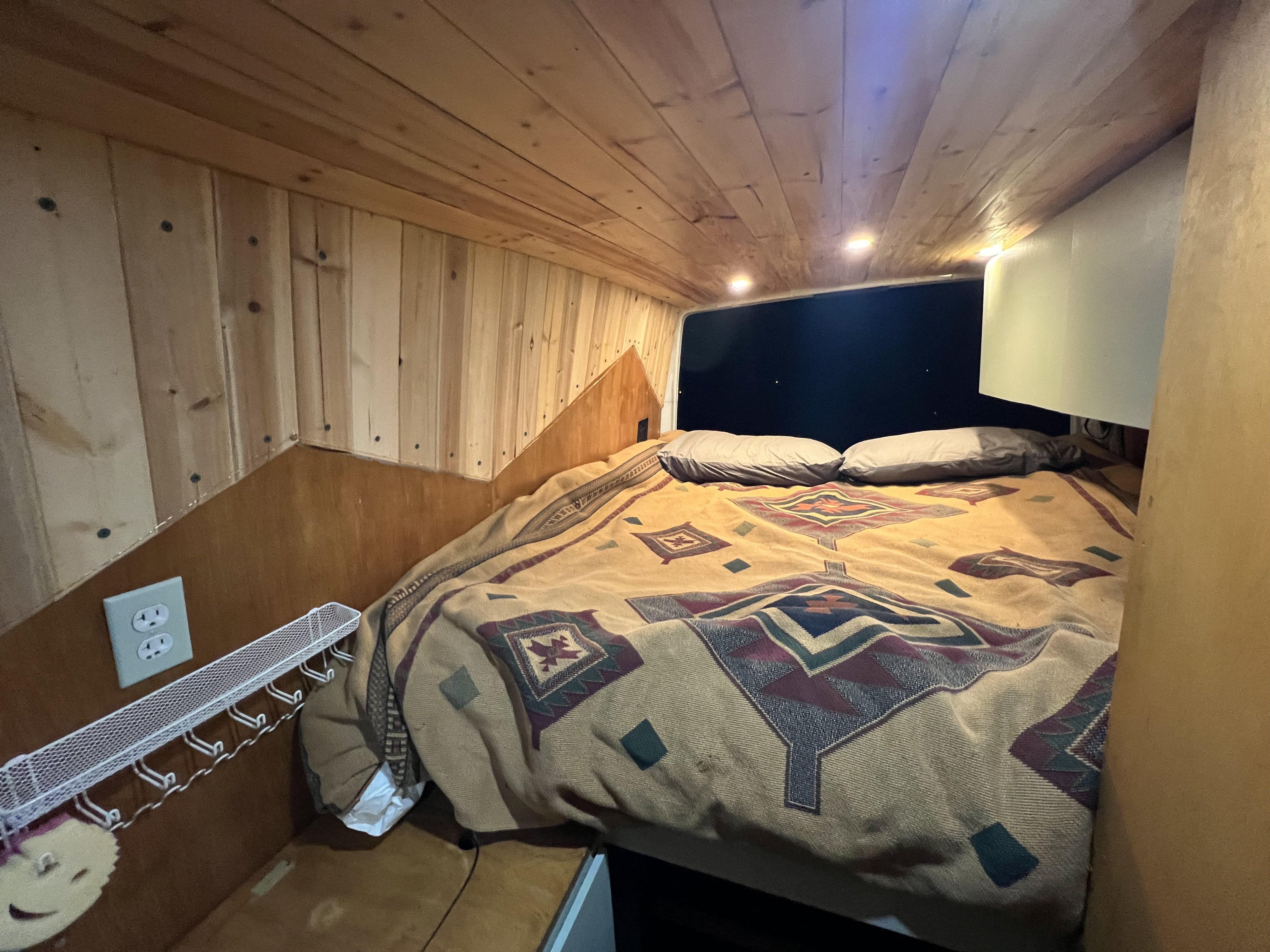 Cozy rv rental bed with patterned blanket, wooden walls/ceiling, recessed lights, and outlet. Ideal camper van sleeping area.. Freightliner Sprinter 2006