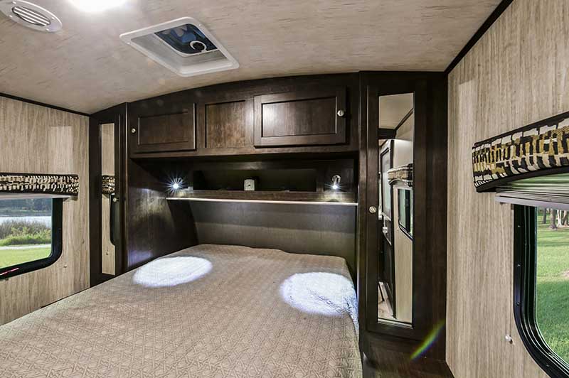RV rental bedroom with overhead dark wood storage, cozy bed lit by bedside lamps, and a window to scenic views.. Cruiser RV Shadow Cruiser 2019