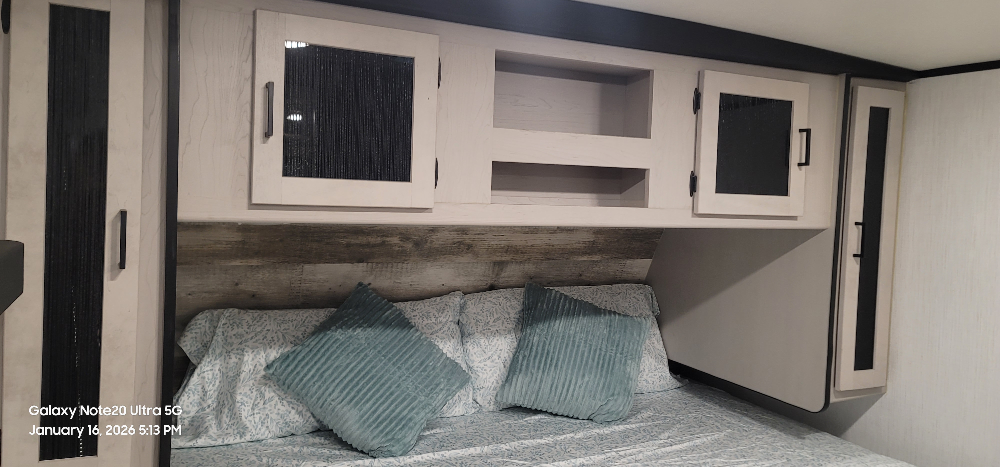 RV rental bedroom showcasing a cozy bed with teal pillows, modern overhead cabinets, and a distressed wood headboard.. Heartland RVs Mallard 2022