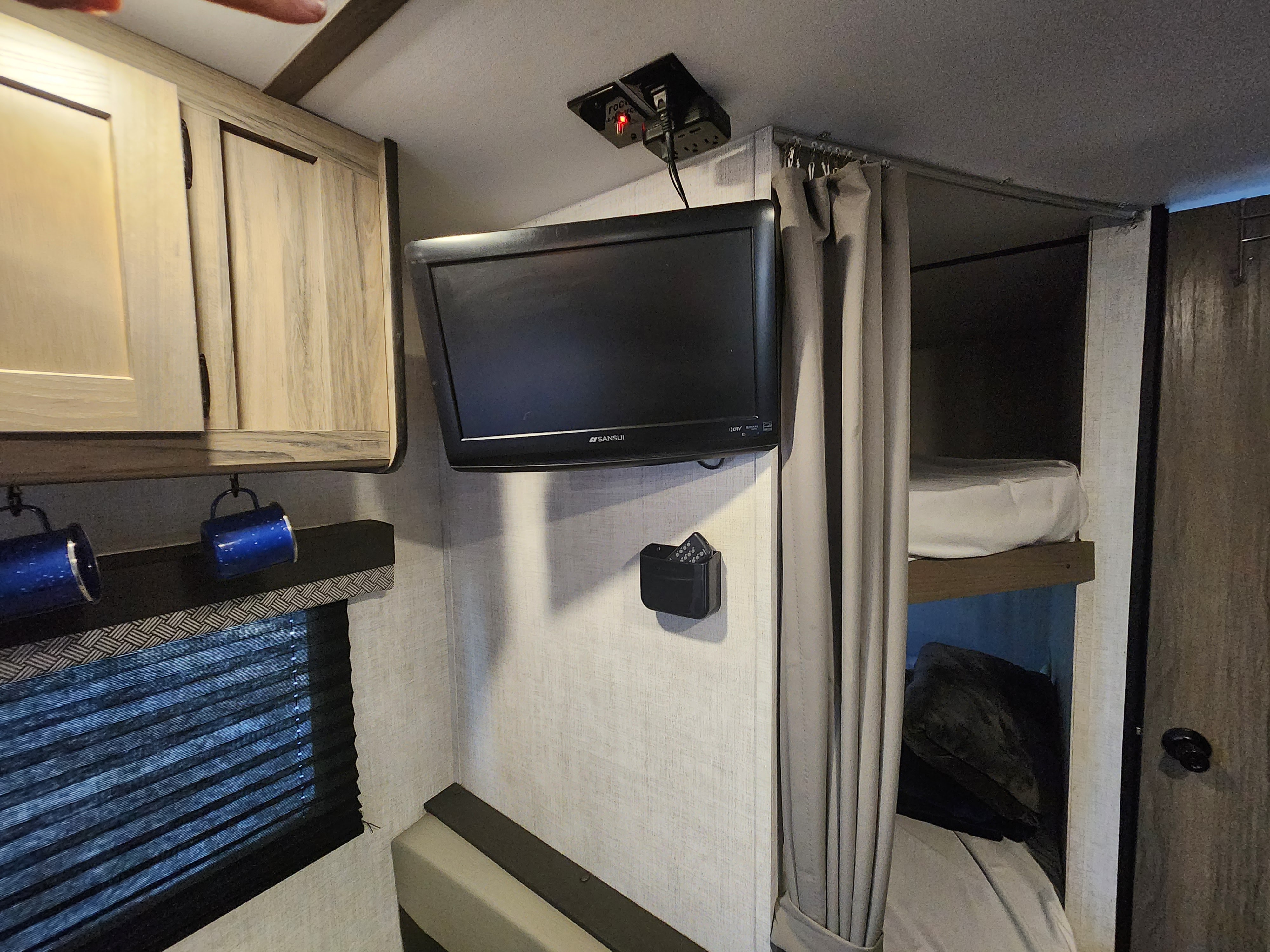 RV rental bunk area with a wall-mounted TV, overhead cabinets, and window blind. A cozy sleeping and entertainment setup.. Gulf Stream Ameri-Lite 2022