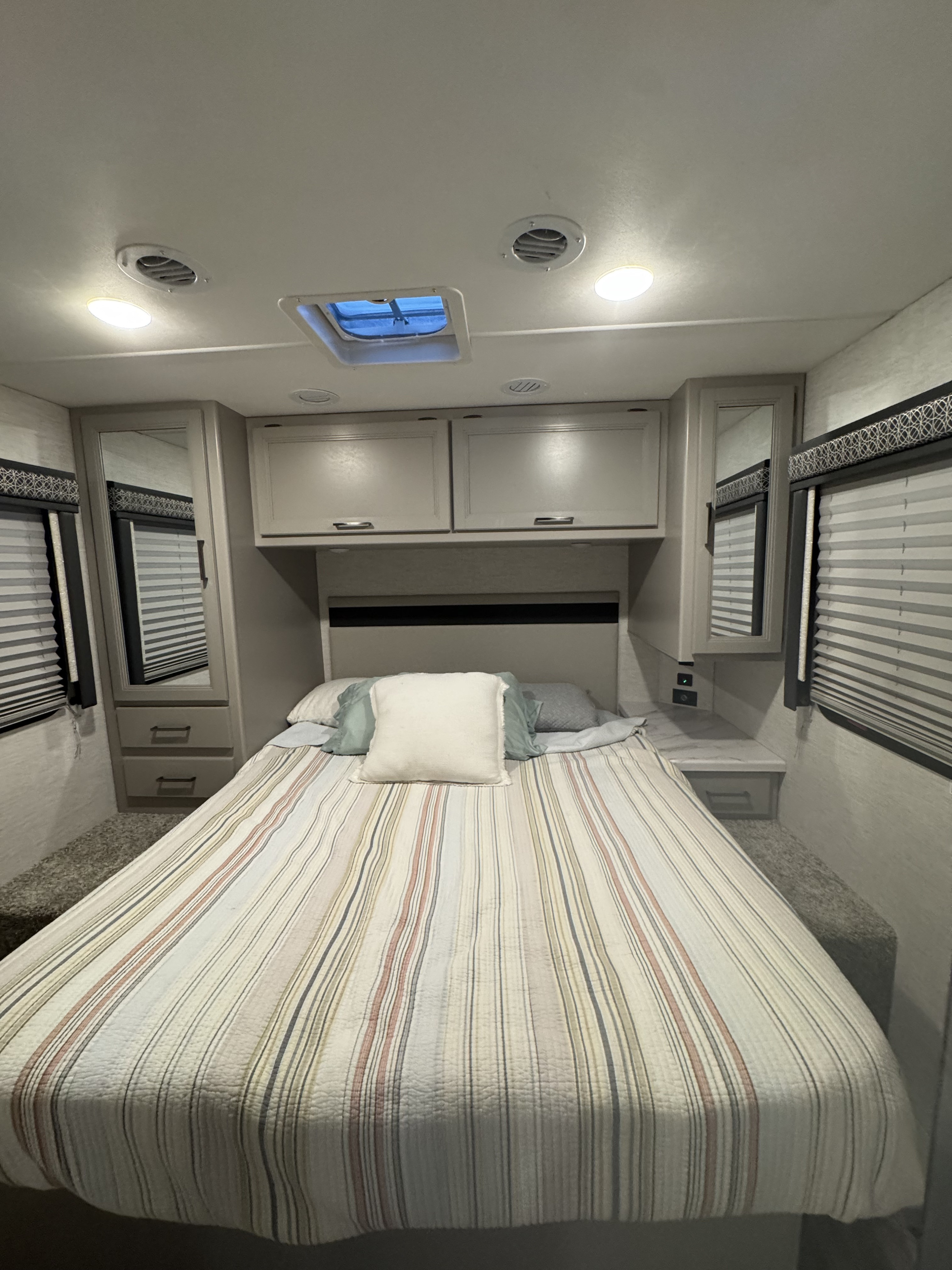Cozy RV rental bedroom featuring a striped bed, overhead cabinets, side mirrors, and blinds. Lights & vents visible.. Thor Quantum SE 2025