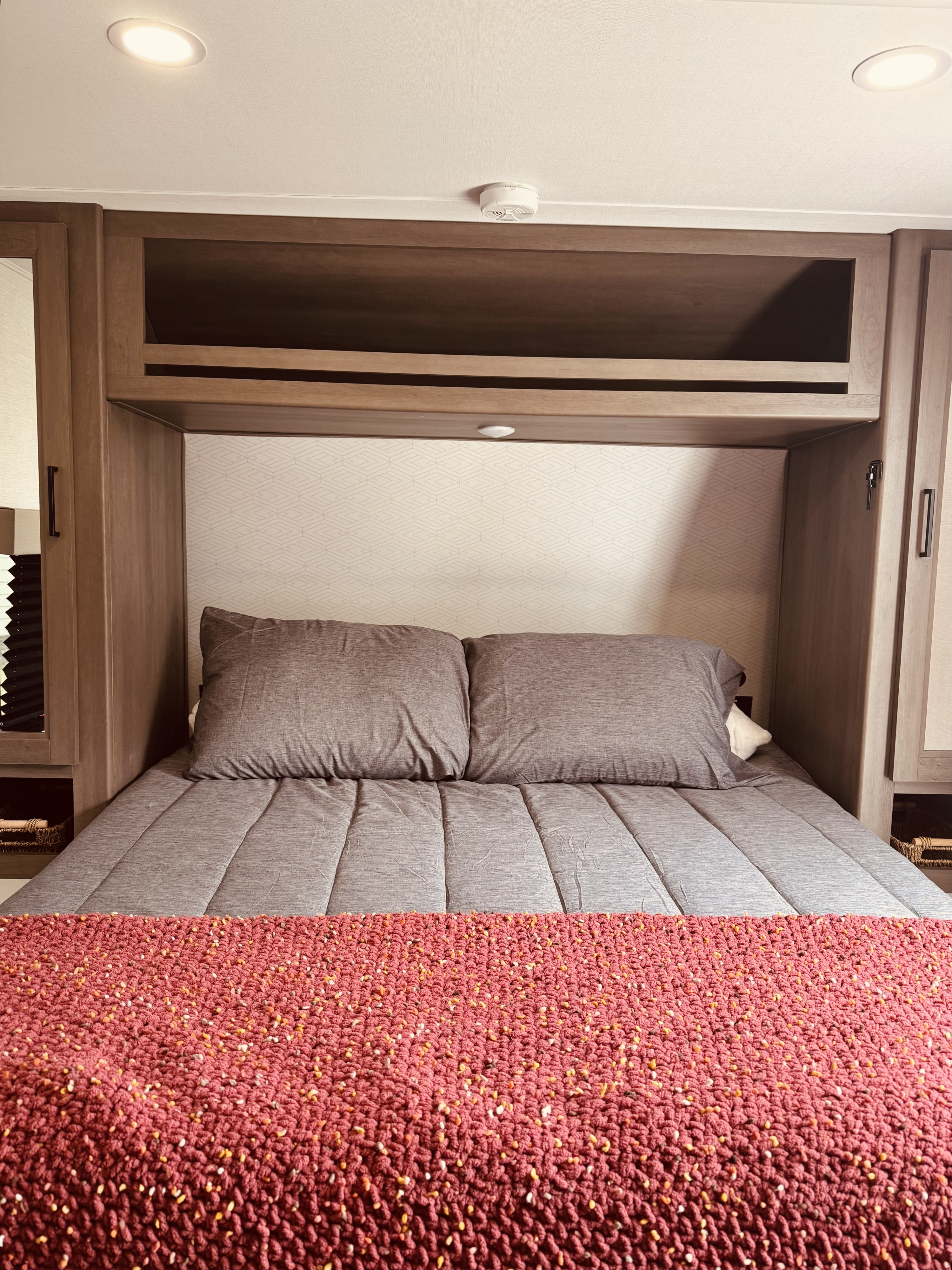 Cozy RV rental bedroom with gray bedding, a textured red blanket, wood cabinetry, and overhead lighting.. Grand Design Transcend One 2025