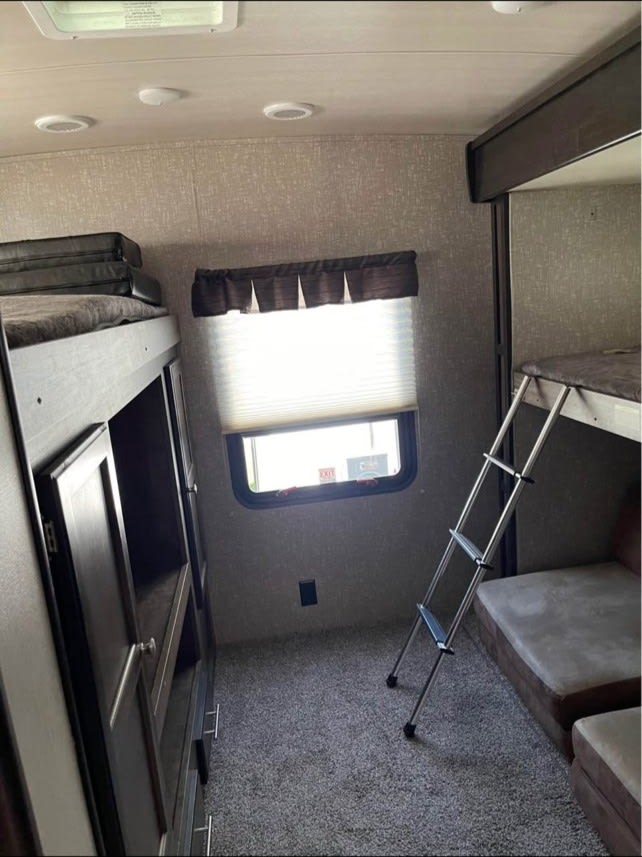 Cozy RV rental interior with bunk beds, a ladder, window with blinds, and built-in storage. Gray carpeted floor.. Heartland RVs Mallard 2020