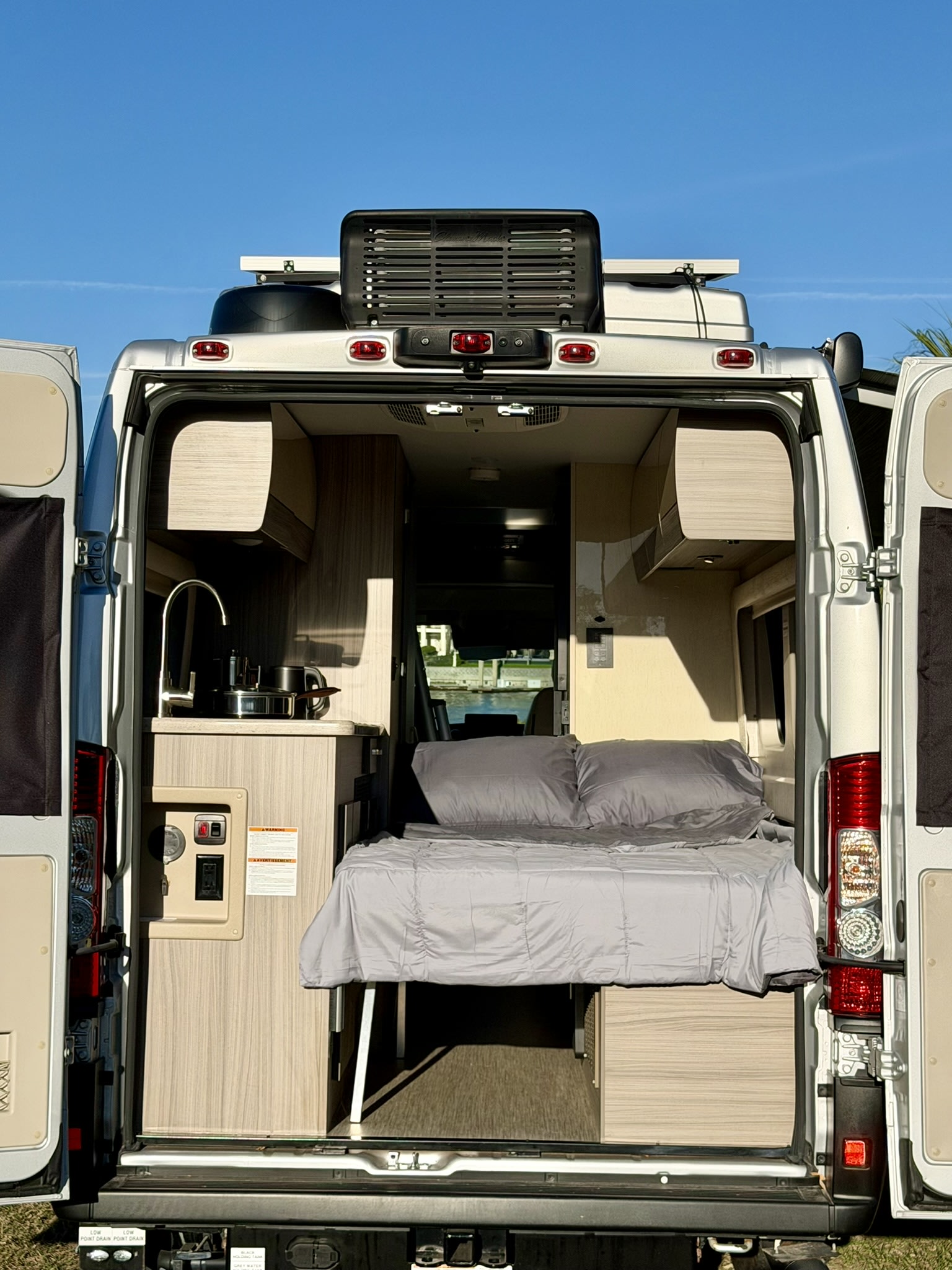 Open rear doors of this RV rental camper van show a compact interior: kitchen with sink, and a neatly made bed, ready for your trip.. Jayco Swift 2024