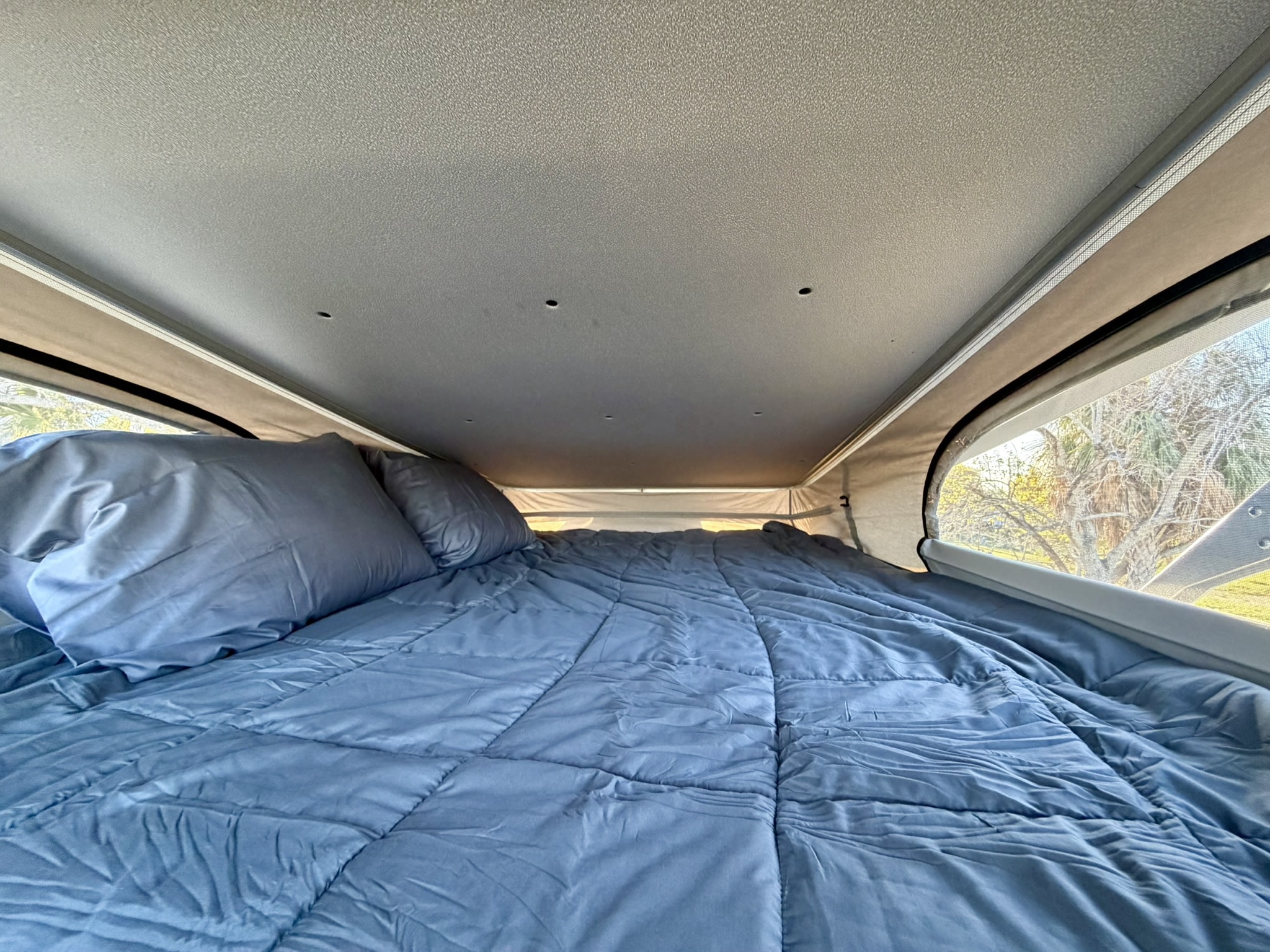 Experience comfort in this RV rental's pop-top bed. A blue comforter and pillows fill the cozy interior, overlooking trees.. Jayco Swift 2024