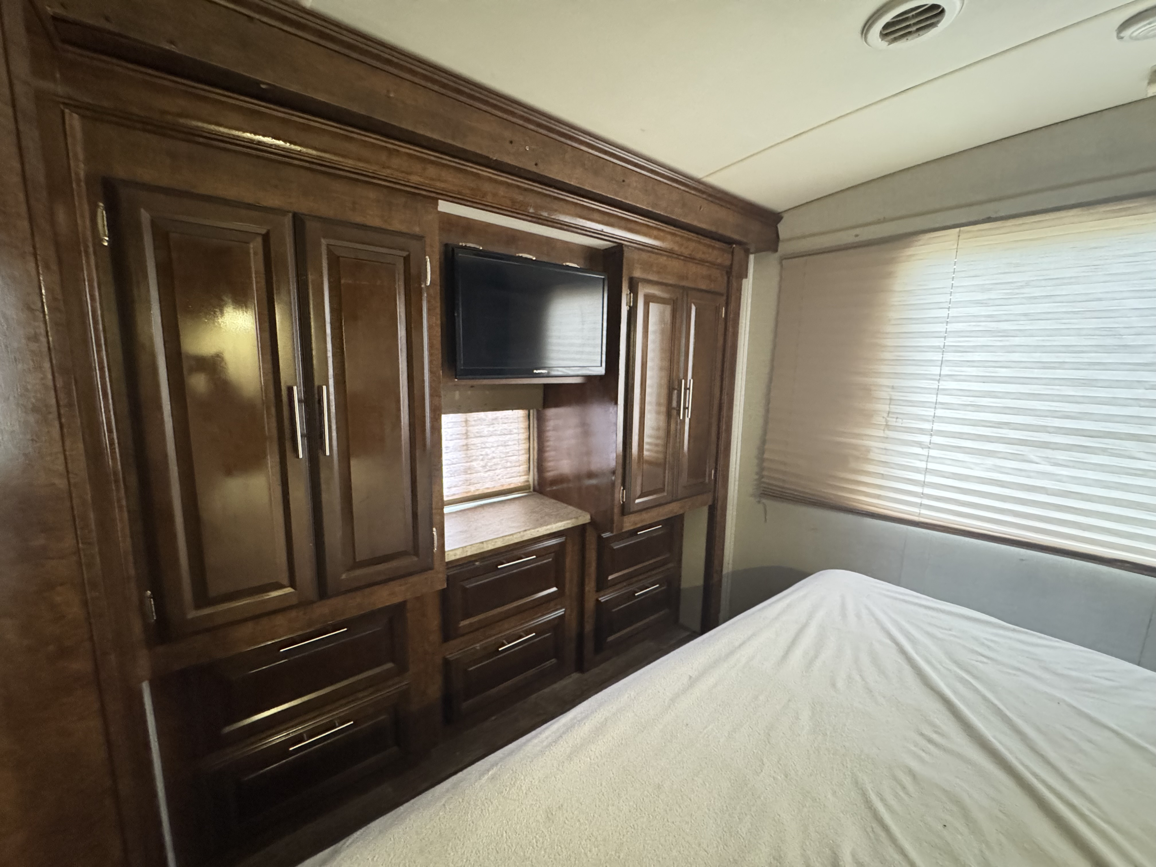 This RV rental's bedroom features dark wood built-in cabinets, a TV, and a window with blinds above the bed.. Forest River FR3 2017