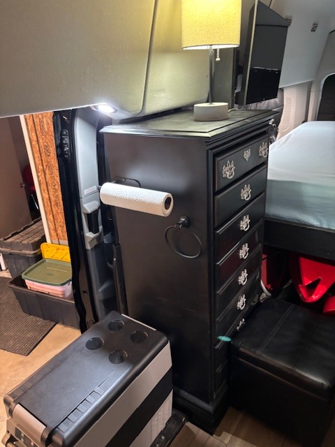 This RV rental features efficient van life design: a tall dresser with a lamp, TV, cooler, and ottoman provides compact storage.. Ford Transit 2019