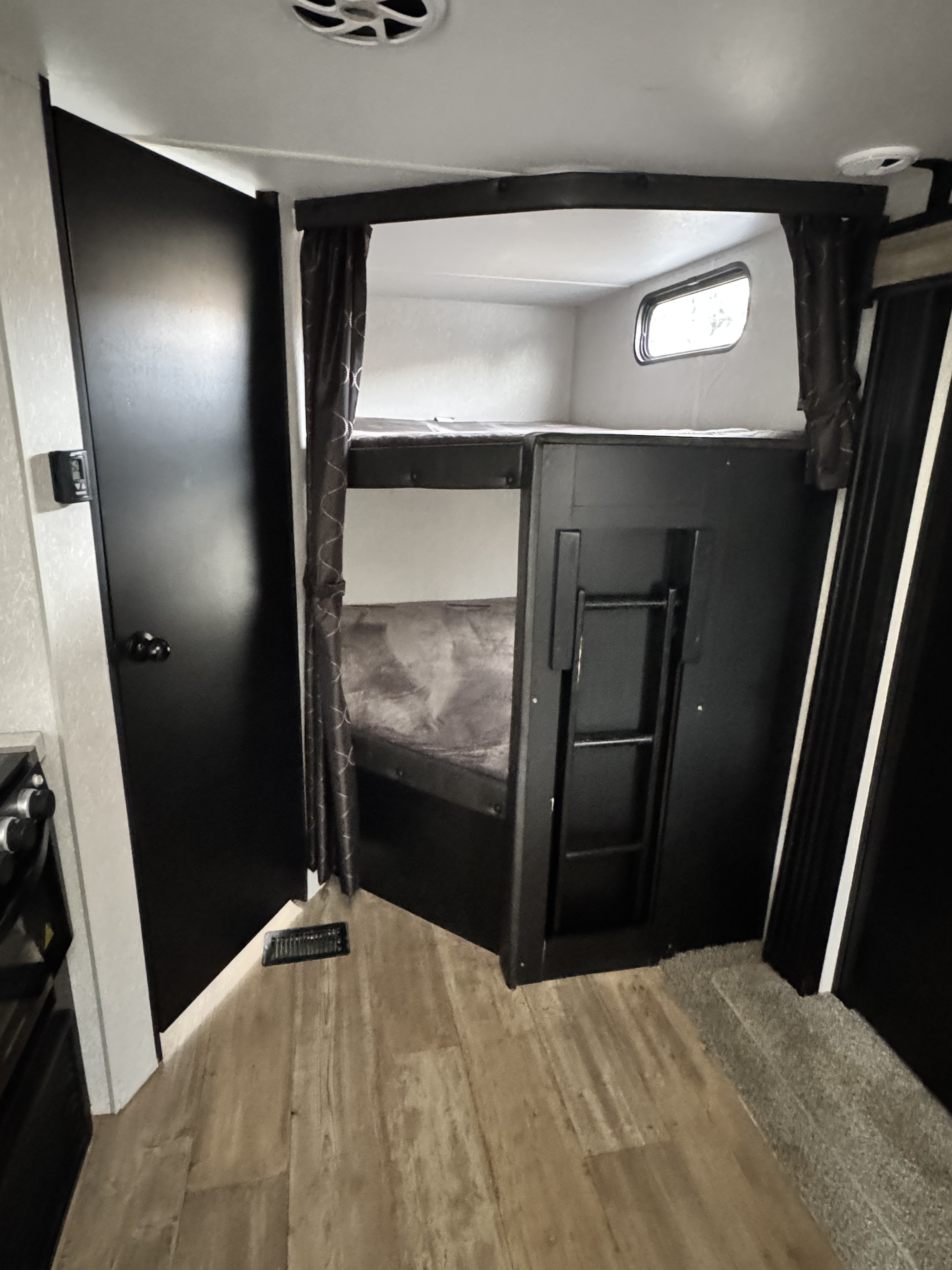 RV rental interior with dark bunk beds, ladder, curtains, and a small window. A black door is visible on the left wall.. Forest River Cherokee 2021