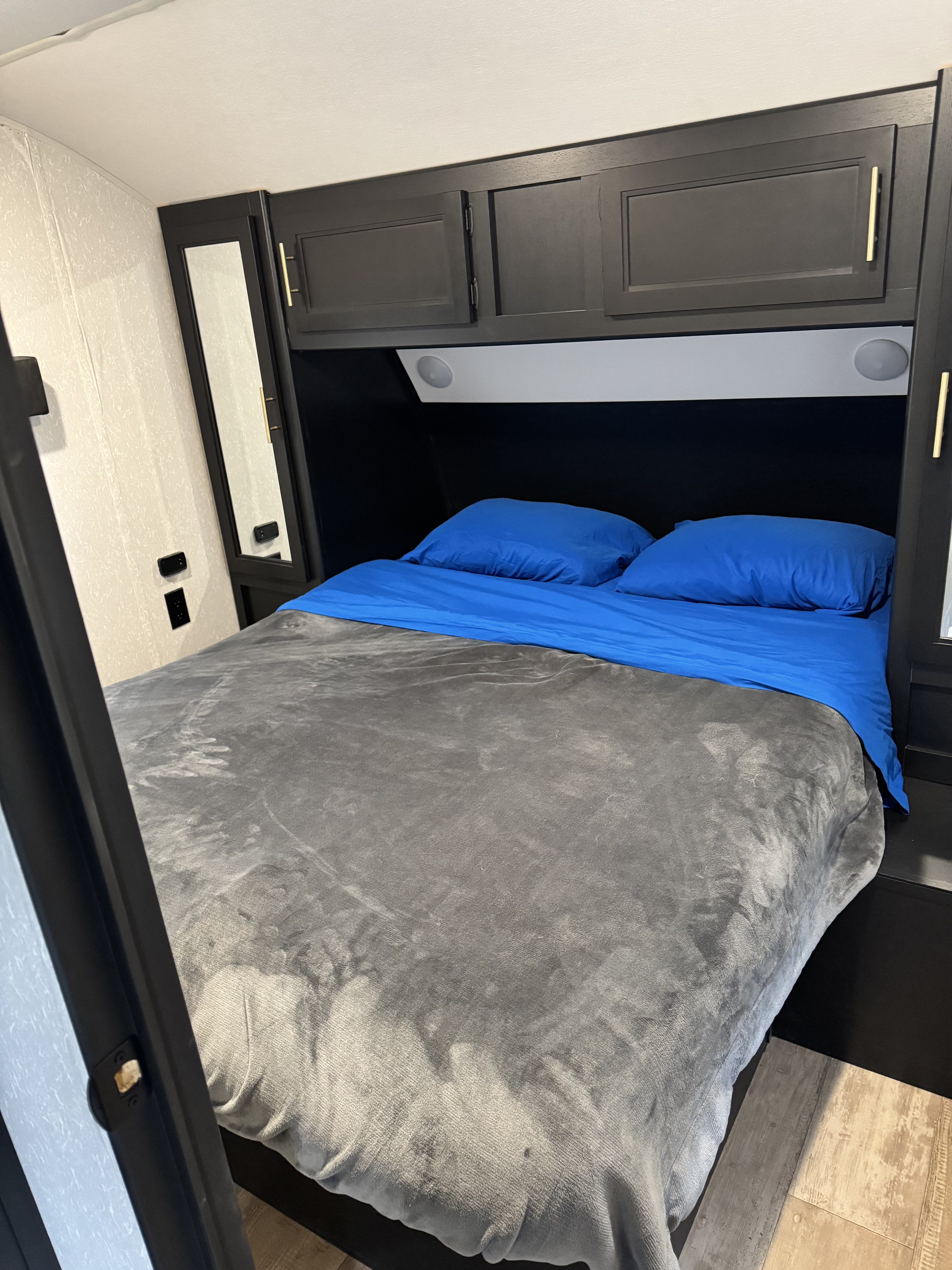 Cozy RV rental bedroom: A comfortable bed with blue linens, grey blanket, dark overhead storage cabinets, and wall outlets.. Forest River Cherokee 2021