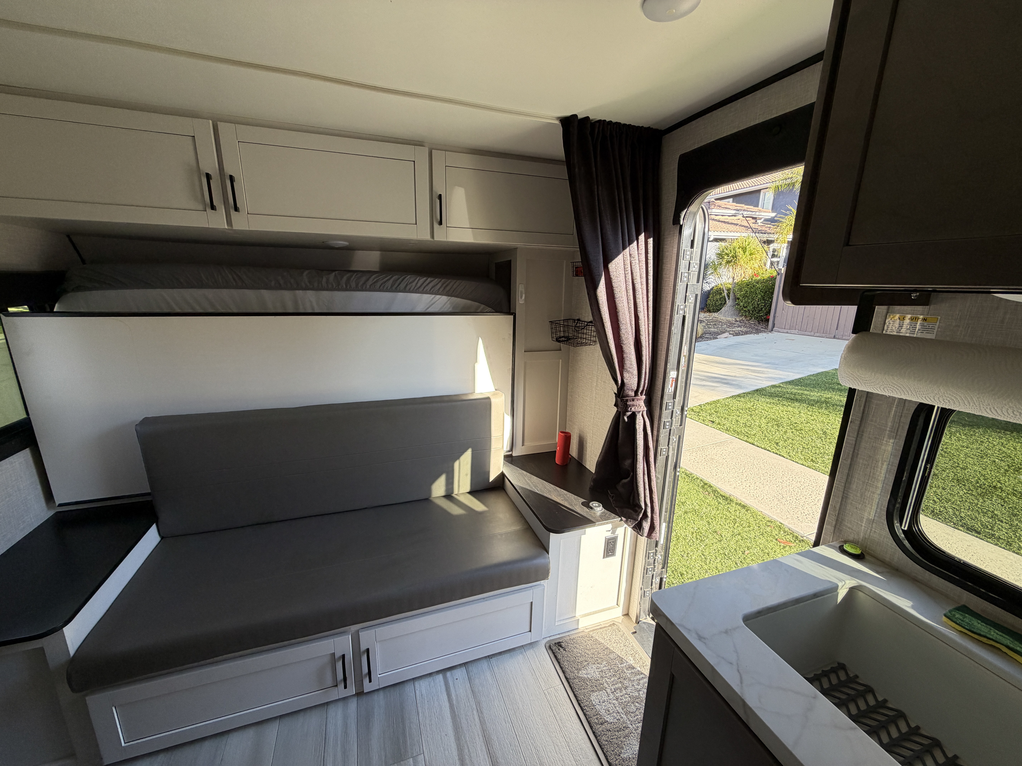 Cozy RV rental interior: sofa seating, an upper bunk, and a compact kitchen area. Bright sunlight streams through the open door.. Autumn Ridge 188BHS 2025