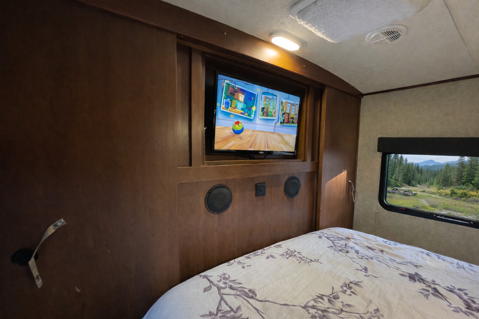 RV rental interior: Bedroom with TV, speakers, and a scenic window showcasing a forest and mountains.. Forest River Palomino Solaire 2020