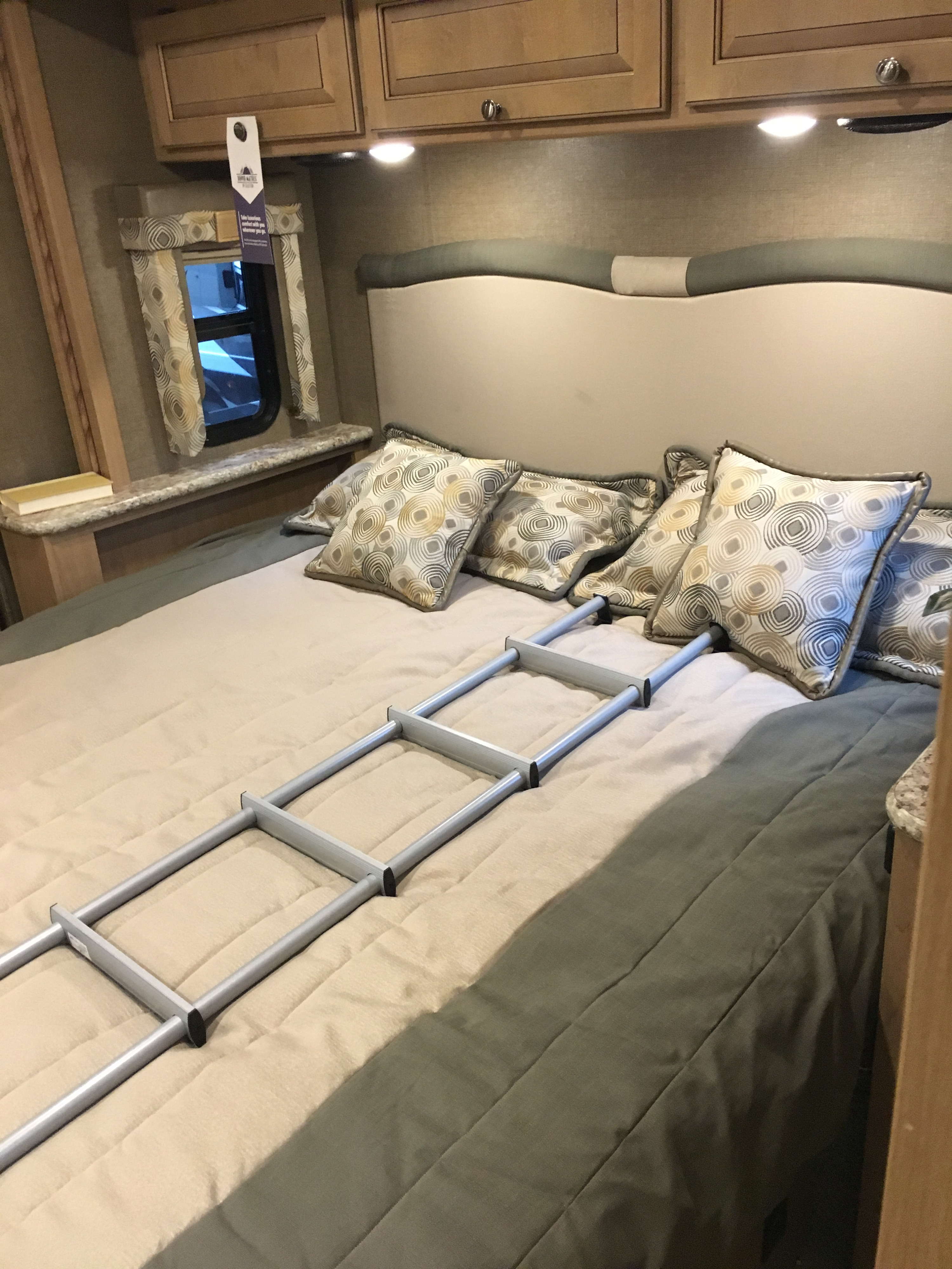 RV rental bedroom interior featuring a made bed with decorative pillows, overhead cabinets, a window, and a ladder.. Thor Windsport 2018