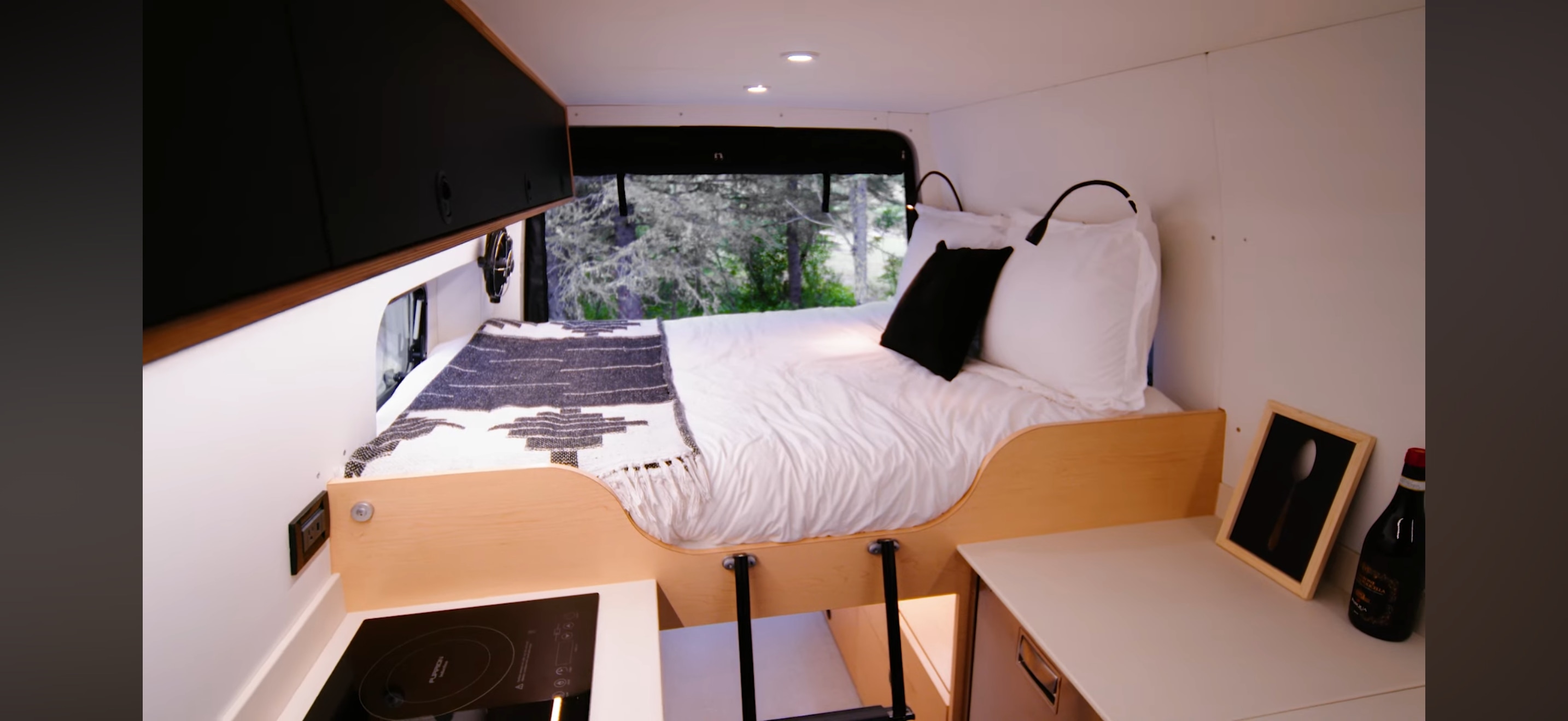 RV rental interior: cozy bed with patterned blanket, compact kitchen, and storage. Outside, lush trees are visible.. MERCEDEZ SPRINTER 17) 4X4 2021