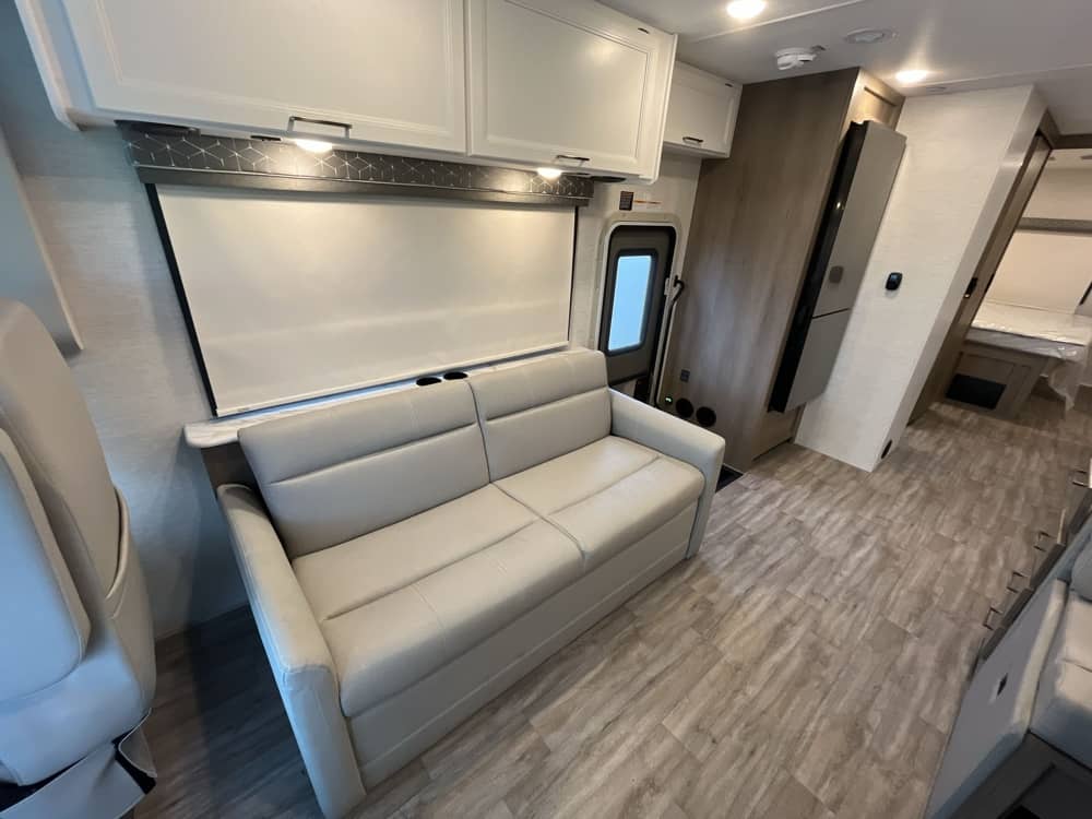 This bright RV rental interior offers a comfortable light-colored sofa, window shade, and modern wood-look flooring leading to a bedroom.. Thor Freedom Traveler 2025