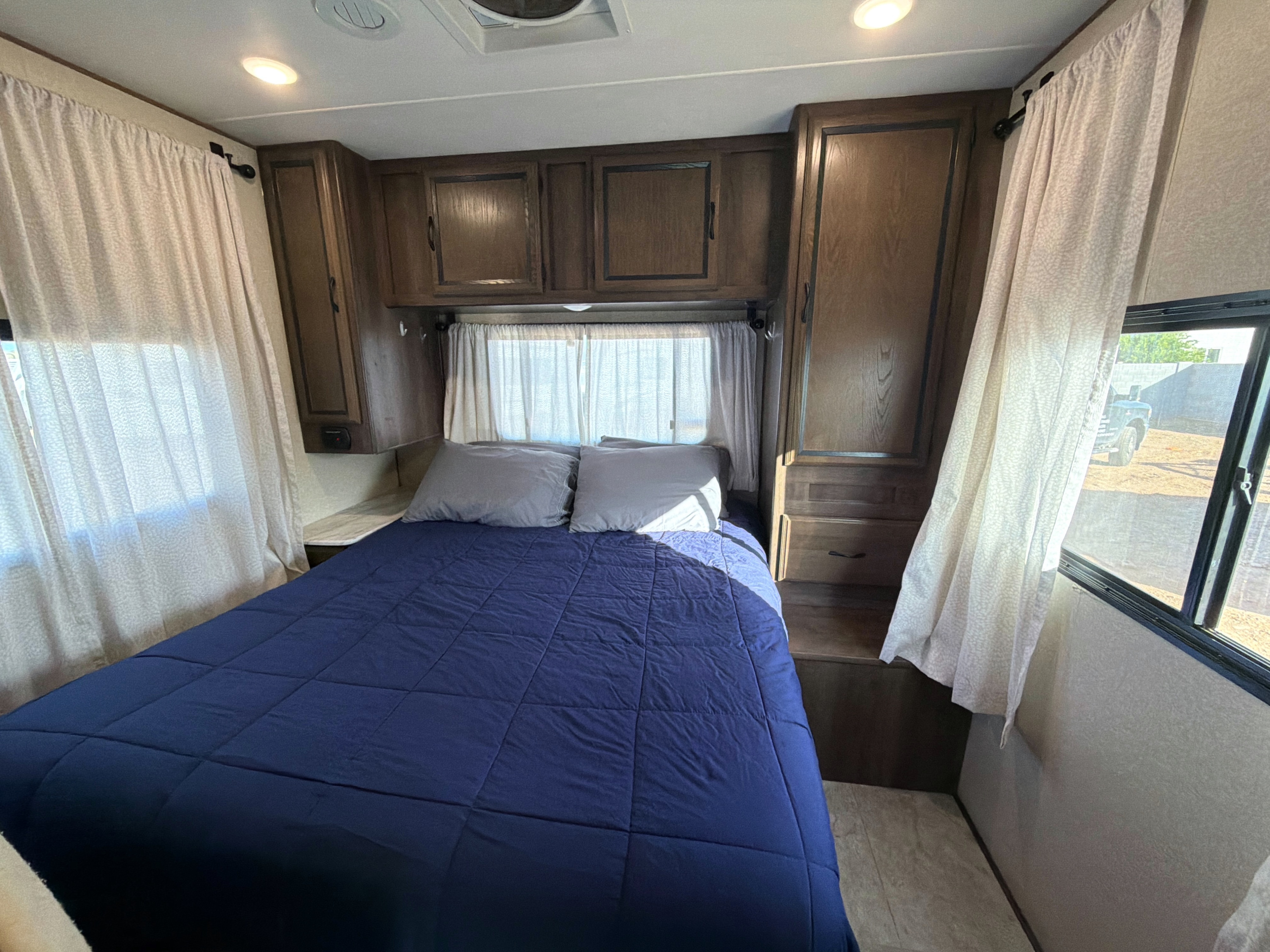 Cozy RV rental bedroom featuring a bed with a blue comforter, light-colored patterned curtains, and wooden cabinets.. Forest River Coachmen Freelander 2018