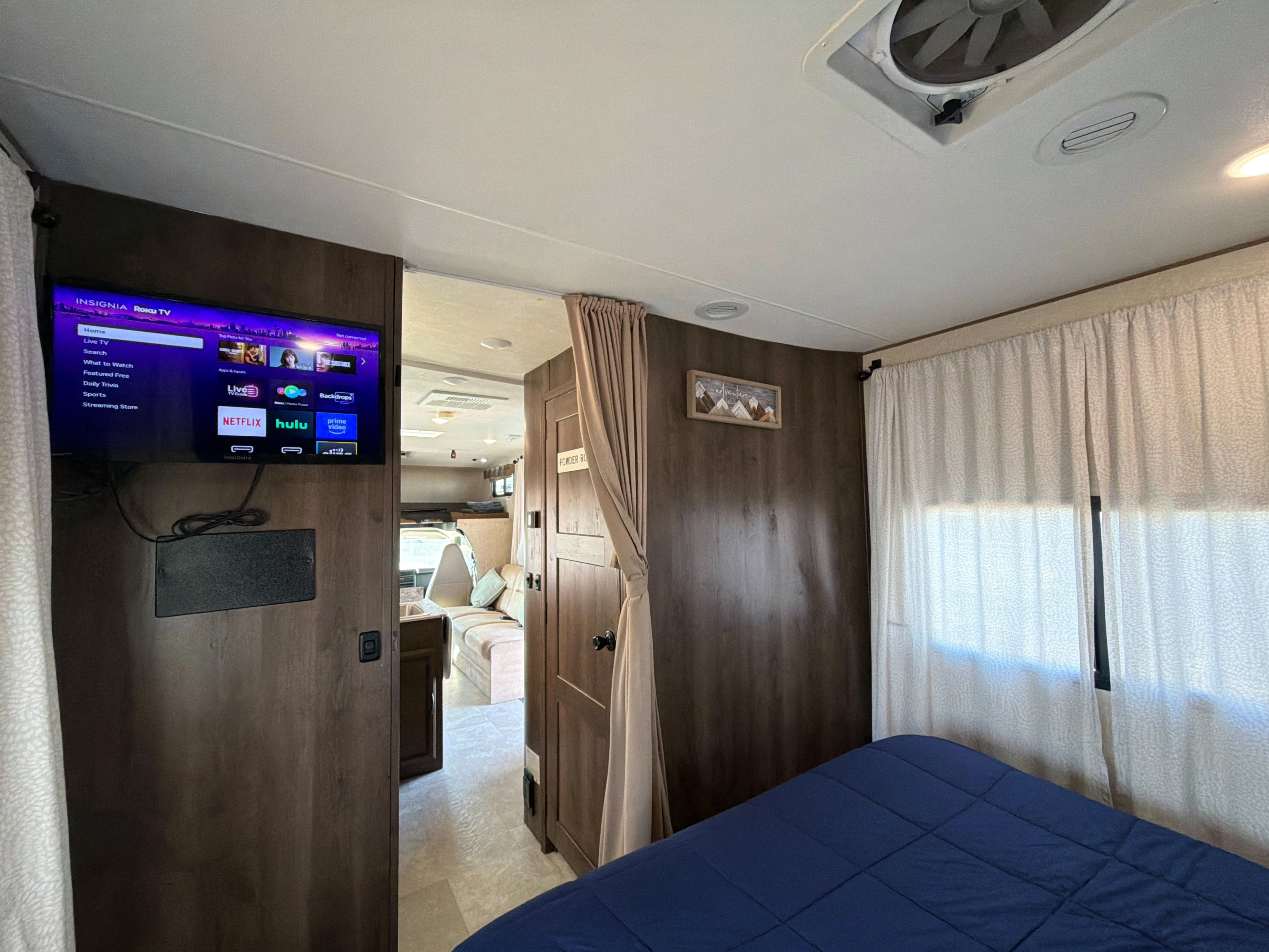 Interior of an RV rental bedroom showing a mounted Roku TV, blue bed, and privacy curtains, with a view into the living area.. Forest River Coachmen Freelander 2018