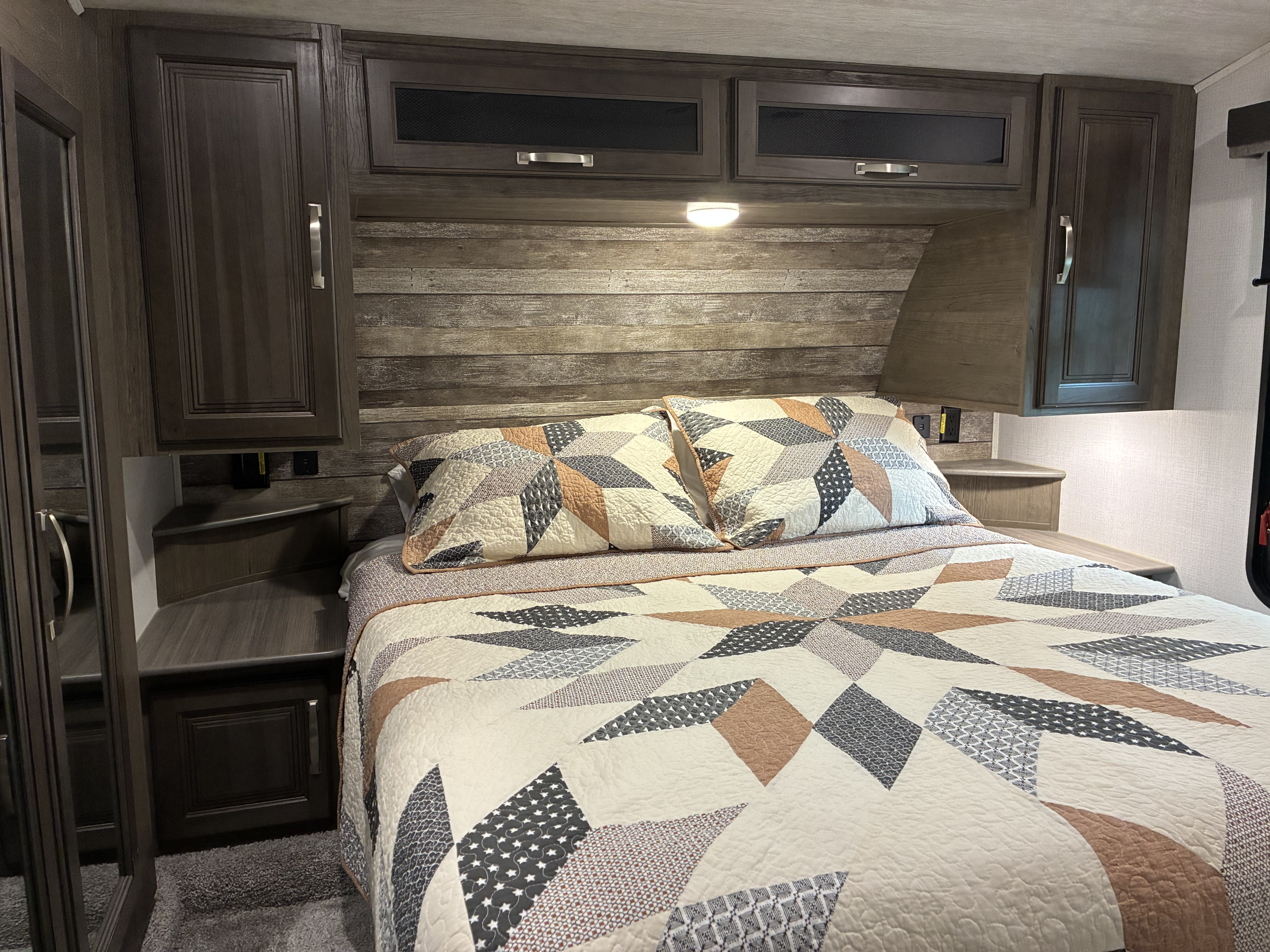 RV rental bedroom featuring a comfortable bed with patterned bedding, overhead cabinets, and a rustic wood headboard.. Keystone RV Cougar 2022
