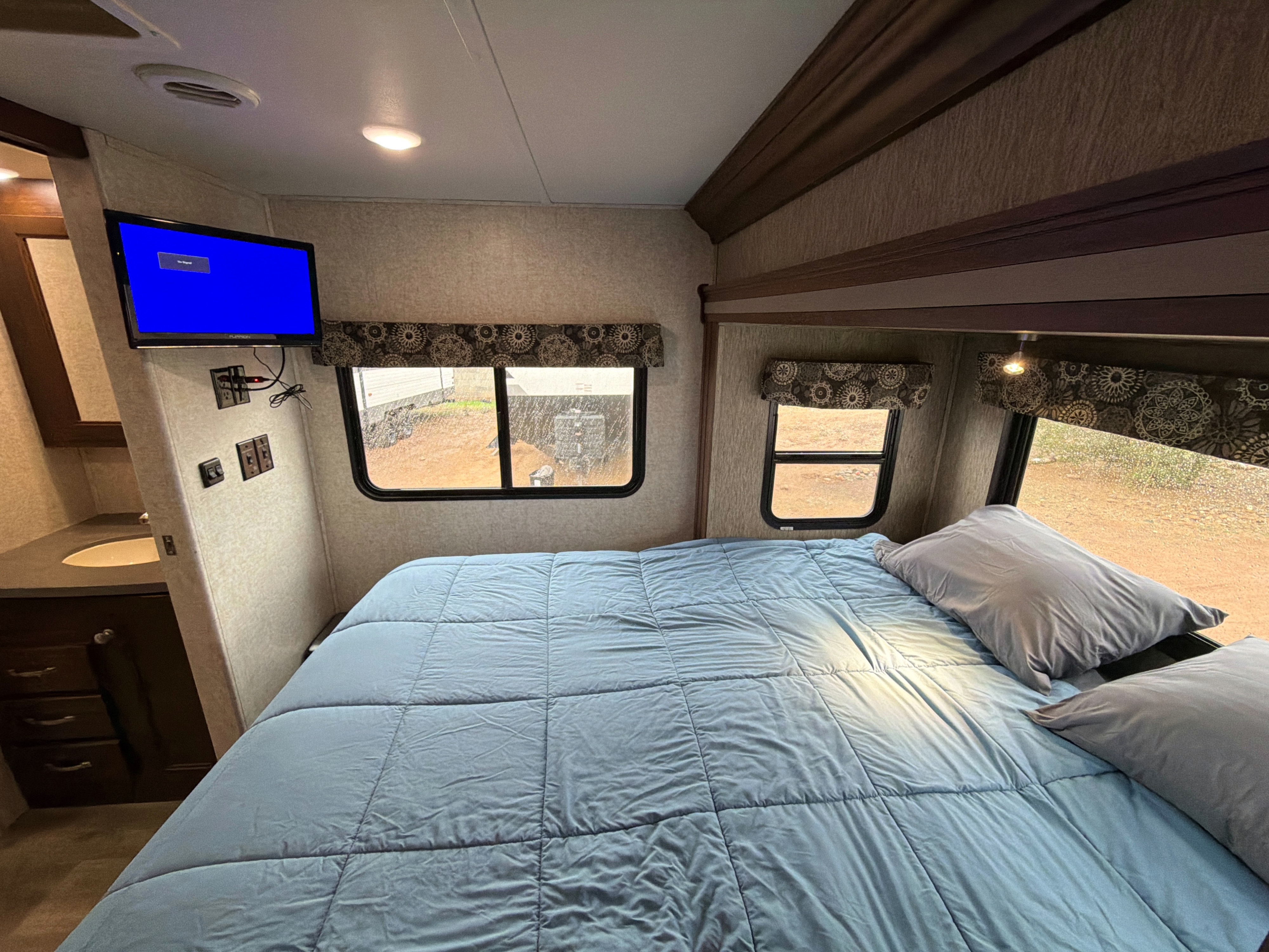 Comfortable RV rental bedroom featuring a blue bed, wall-mounted TV, vanity, and large windows revealing the outdoors.. Forest River Sunseeker 2017