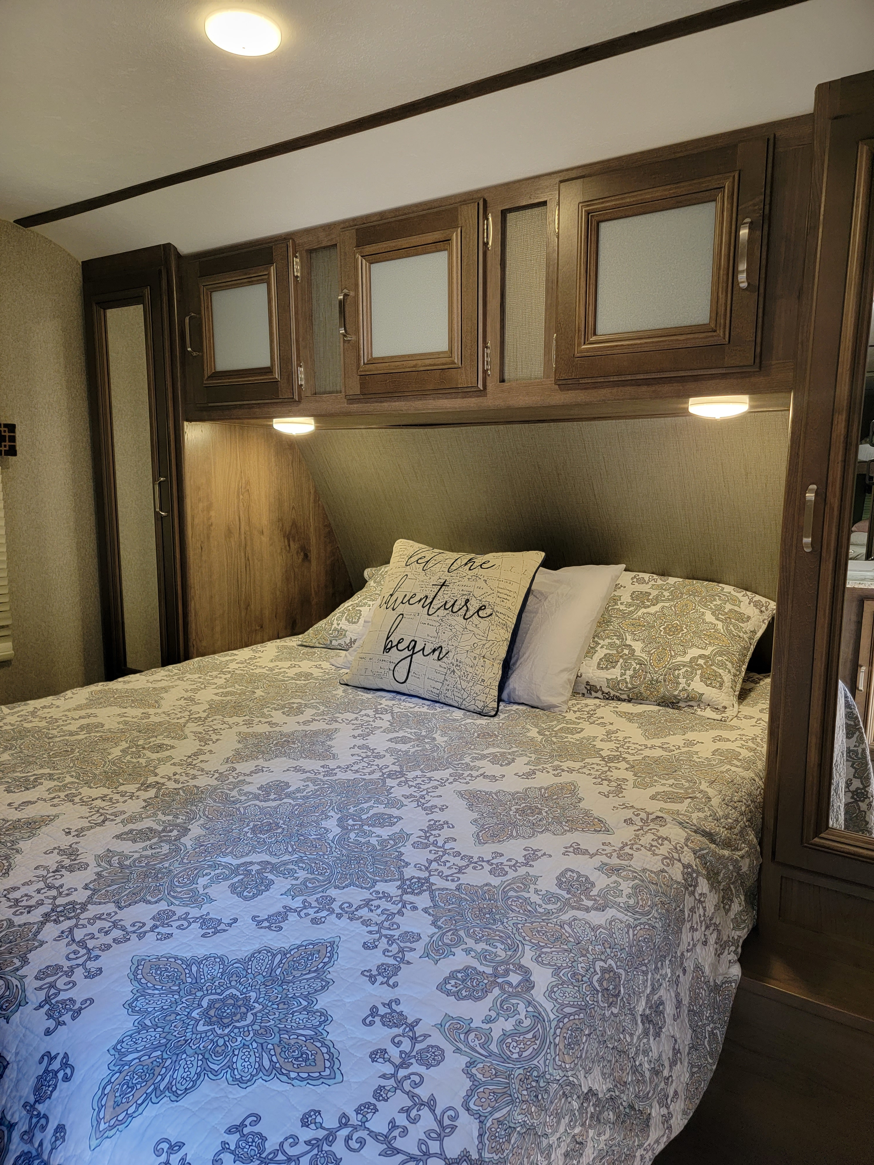 Cozy RV rental bedroom featuring a patterned bed, overhead wood cabinets, and integrated bedside lighting for travel adventures.. Keystone RV Passport Grand Touring 2018