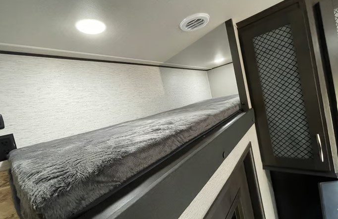 Cozy RV rental bunk bed with gray fluffy mattress, overhead lights, and a mesh cabinet in a modern interior.. Grand Design Momentum M-Class Toy Hauler 2023