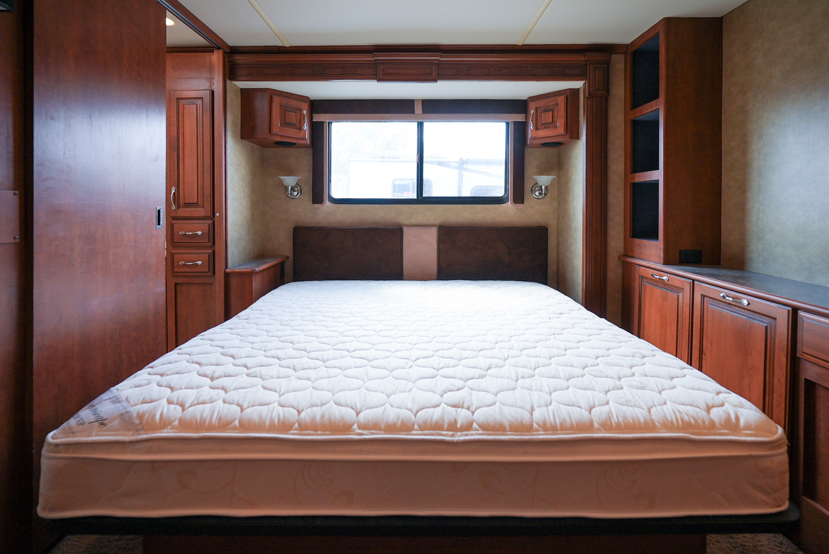 This RV rental bedroom showcases a large, quilted mattress, dark wood cabinetry, a window, and wall sconces.. Southwind Southwind Motorhome 2013