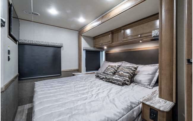 RV rental bedroom featuring a spacious bed, overhead cabinets, roller blinds, and a TV for ultimate comfort.. Holiday Rambler Admiral 2019
