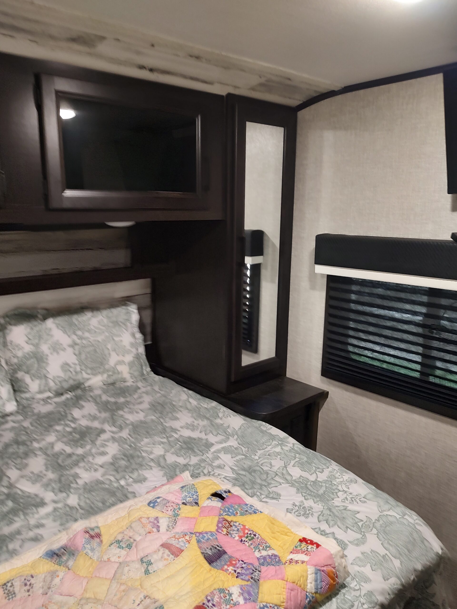 RV rental bedroom: Cozy bed with floral linens, colorful quilt, mounted TV, mirror, dark wood cabinets, and window with blinds.. Jayco Jay Feather 2022