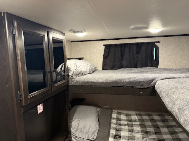 Inside an RV rental: a cozy bunk bed area with grey bedding, dark wood cabinet, and overhead lights for comfort.. Jayco Jay Feather 2021
