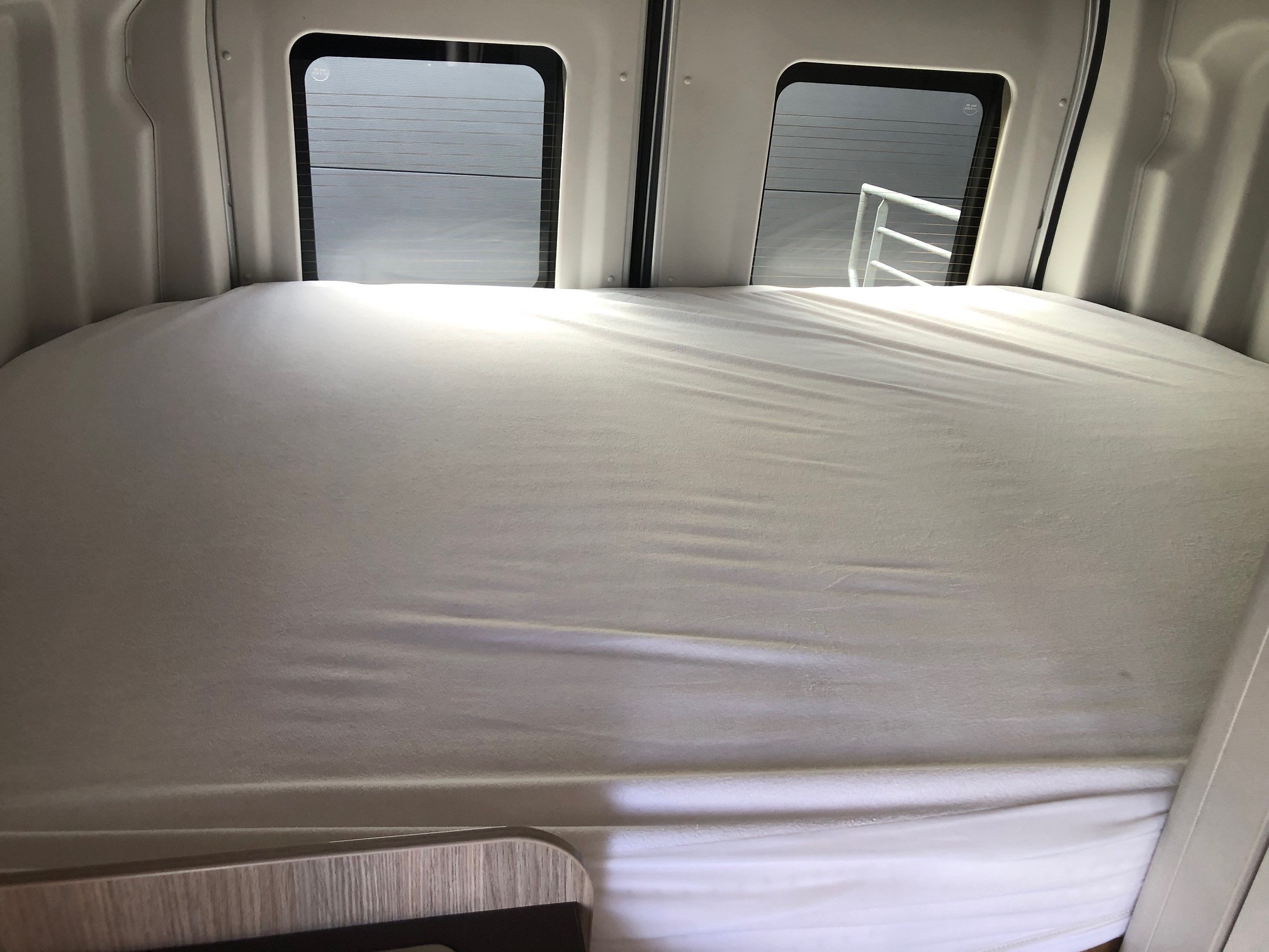 RV rental interior: A bed with white sheets fills the rear, with two windows showing an exterior building and railing.. BURSTNER LINEO C590 GRIS 2023