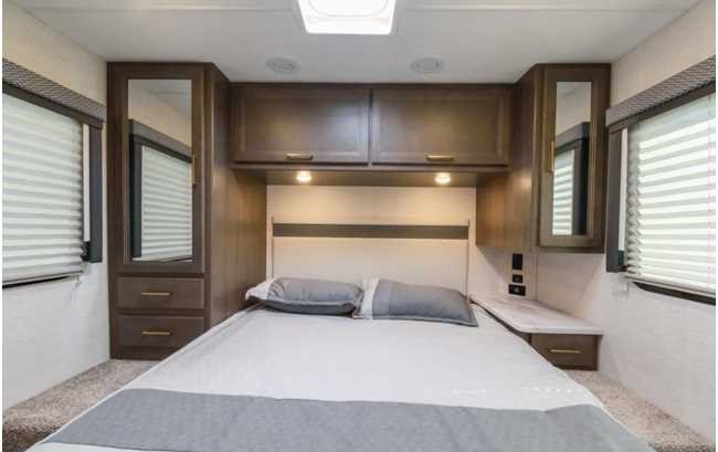 RV rental bedroom with a cozy bed, ample overhead and side storage, reflective cabinet doors, and bright windows with blinds.. Thor Four Winds 2025