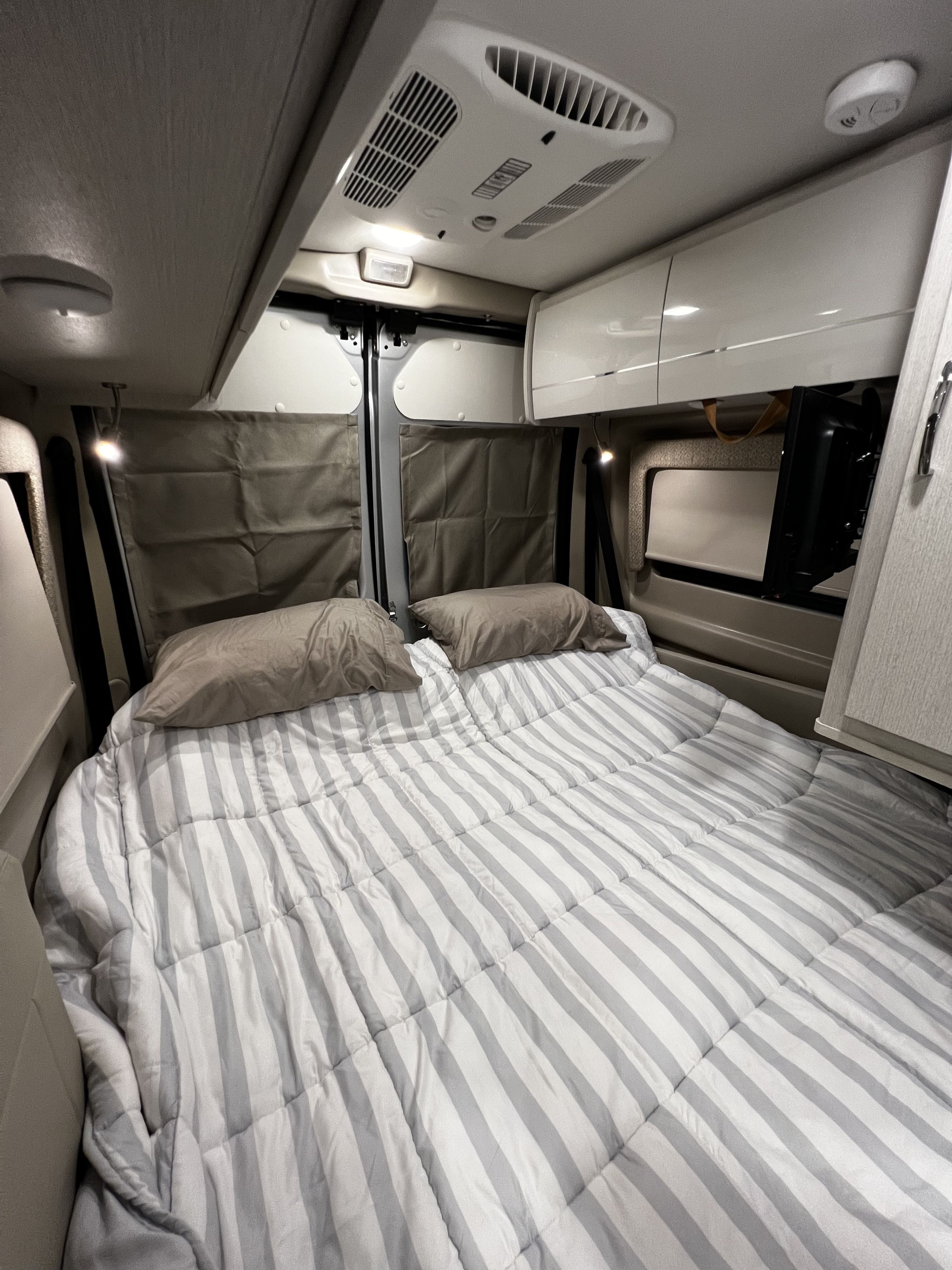 Cozy RV rental bedroom featuring a comfy striped bed, overhead storage, AC, and reading lights for your next adventure.. Thor Tellaro 2022