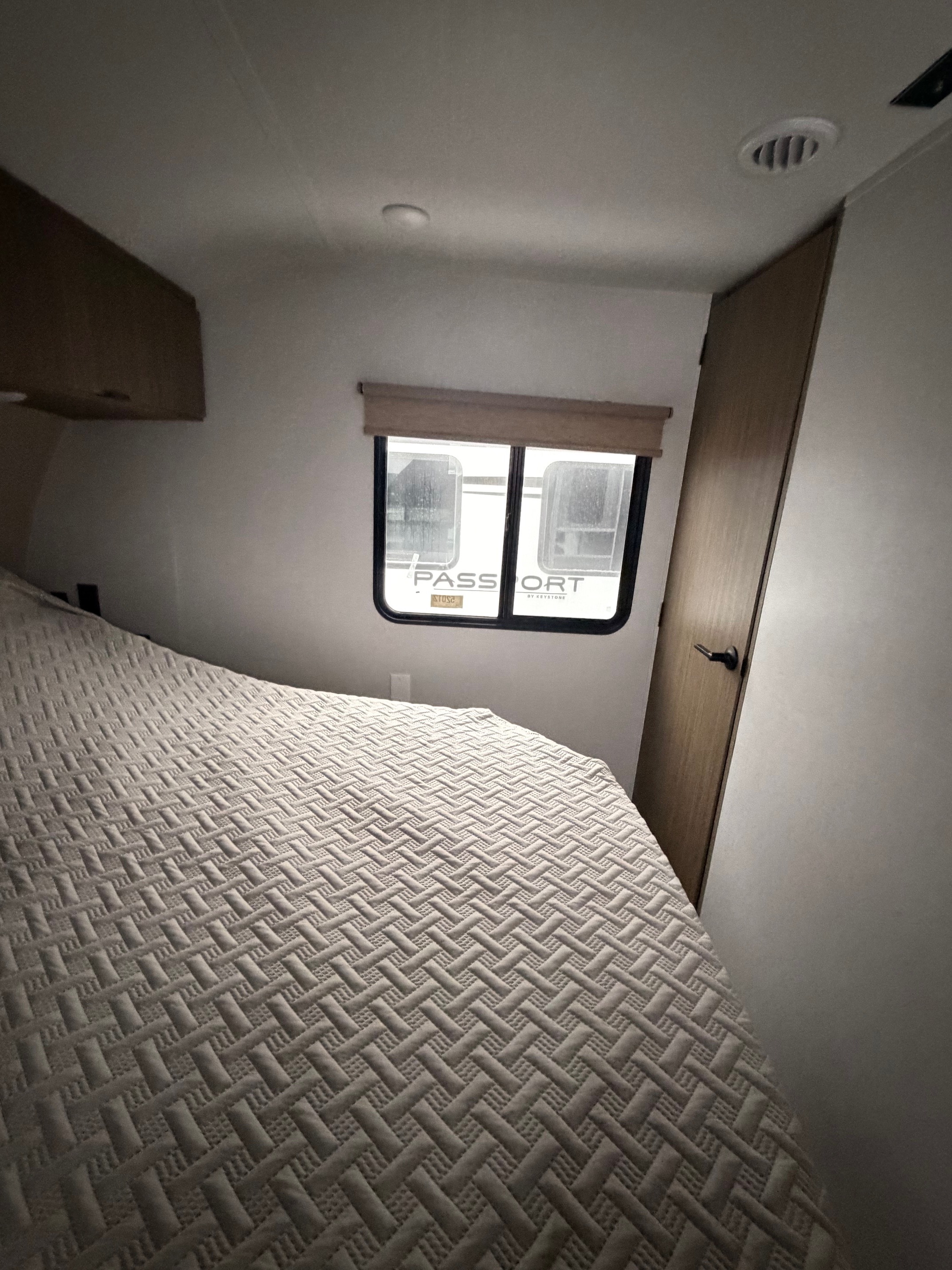 This RV rental bedroom features a comfy patterned bed, window with 'Passport' RV outside, overhead storage, and a door.. Forest River Wildwood 2025