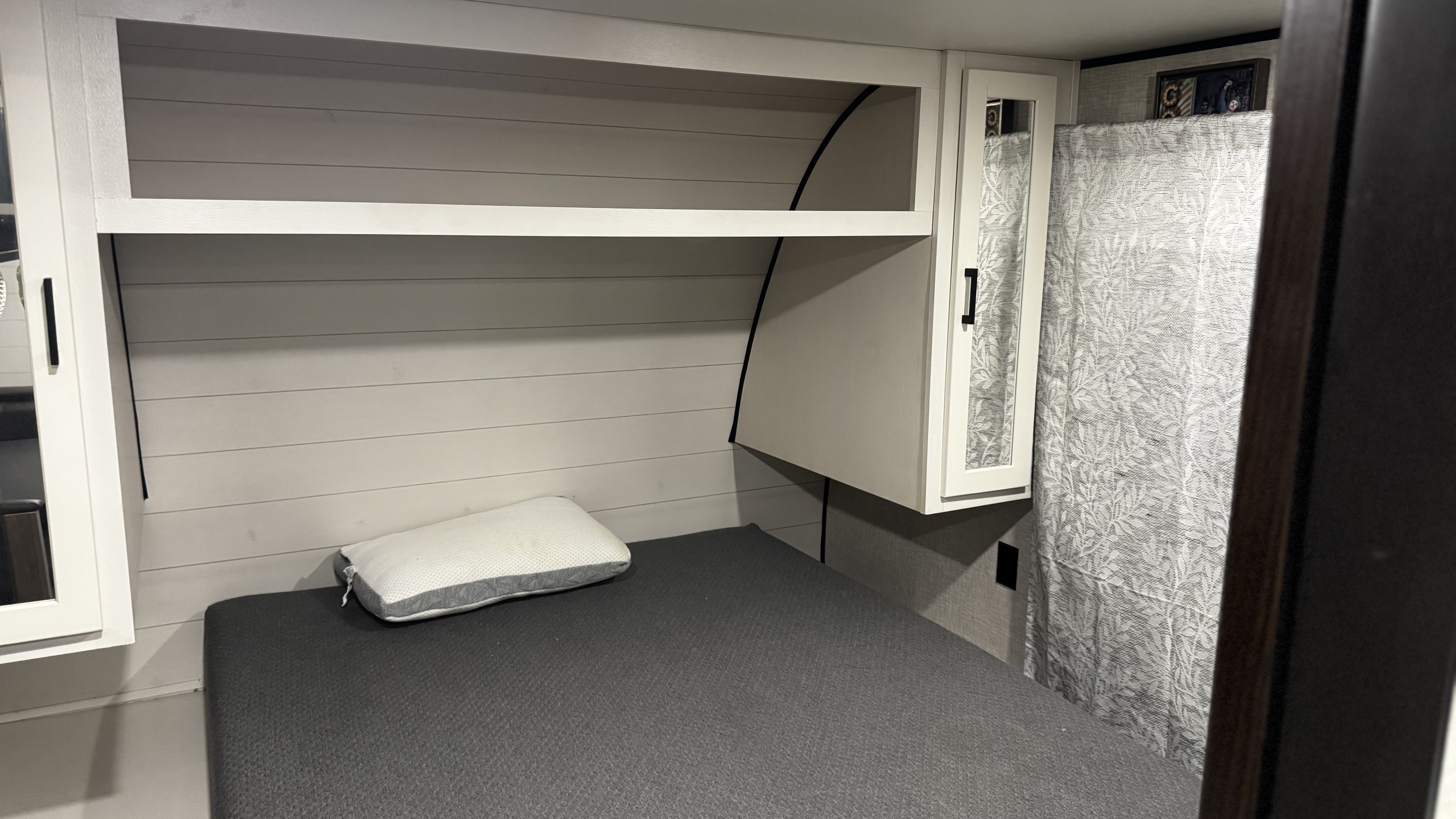 RV rental bedroom: a grey mattress & white pillow under built-in white shelving. Modern, compact interior with patterned curtain.. Jayco Jay Flight Baja Edition 2024