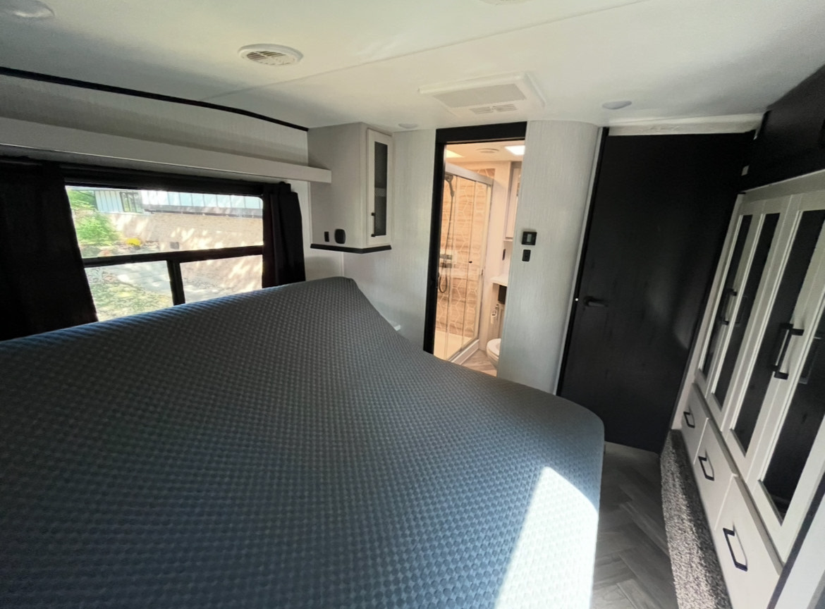 Modern rv rental bedroom features a bed, window, and open bathroom with a shower. Built-in storage on right.. Heartland RVs Cyclone Toy Hauler 2022