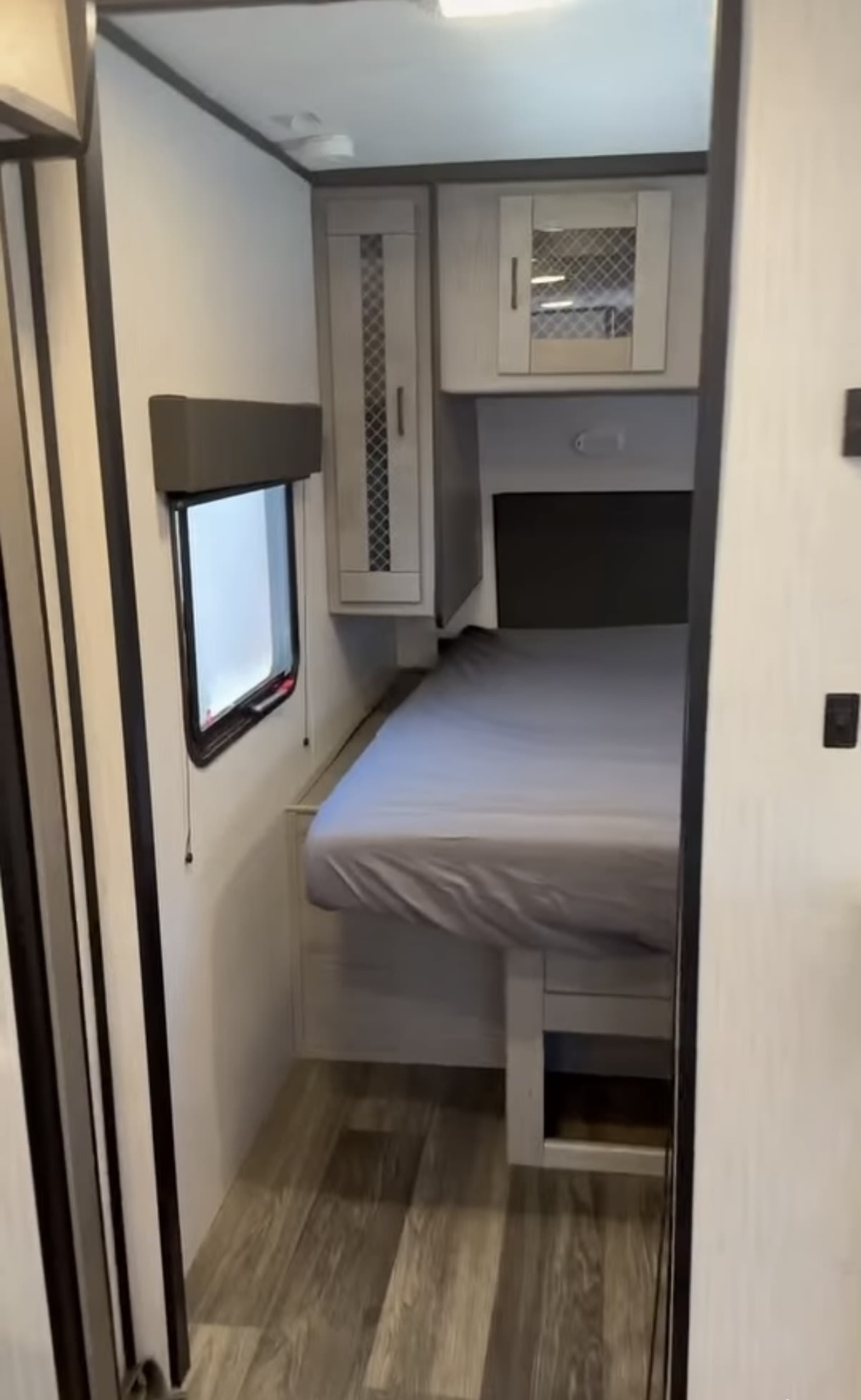 Cozy RV rental bedroom featuring a bed with gray sheets, a window, wood-look floor, and overhead storage cabinets.. Heartland RVs Mallard 2021