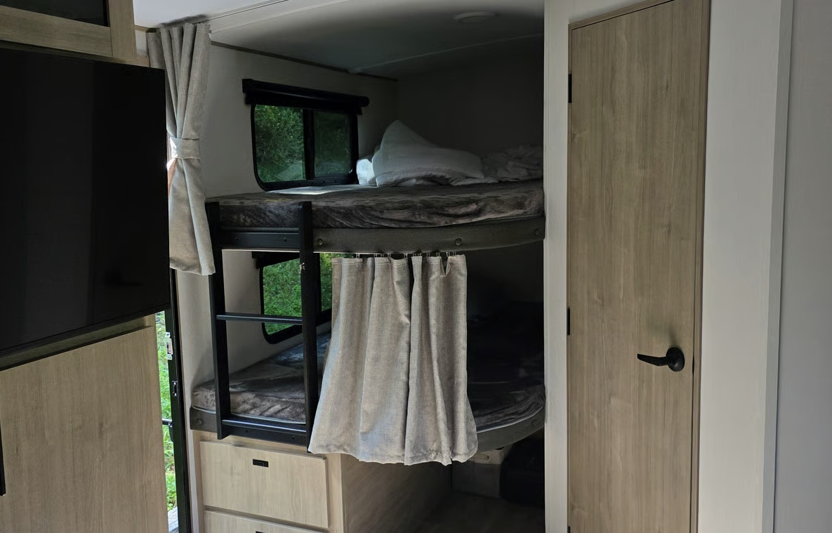 Inside this rv rental, bunk beds with windows and privacy curtains are visible. A TV is mounted left, beside a light-wood door.. Grand Design 2800BH 2026