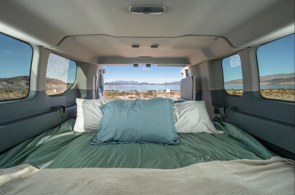 RV rental: Interior of a camper van with a neatly made bed and pillows, looking out to a vast lake and distant mountains.. Ford Transit 2017