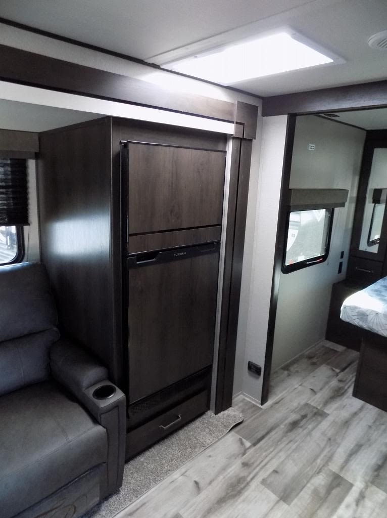 RV rental interior: a dark wood refrigerator unit, comfy gray armchair, and a glimpse into the bedroom.. Grand Design Transend Xplor 2024