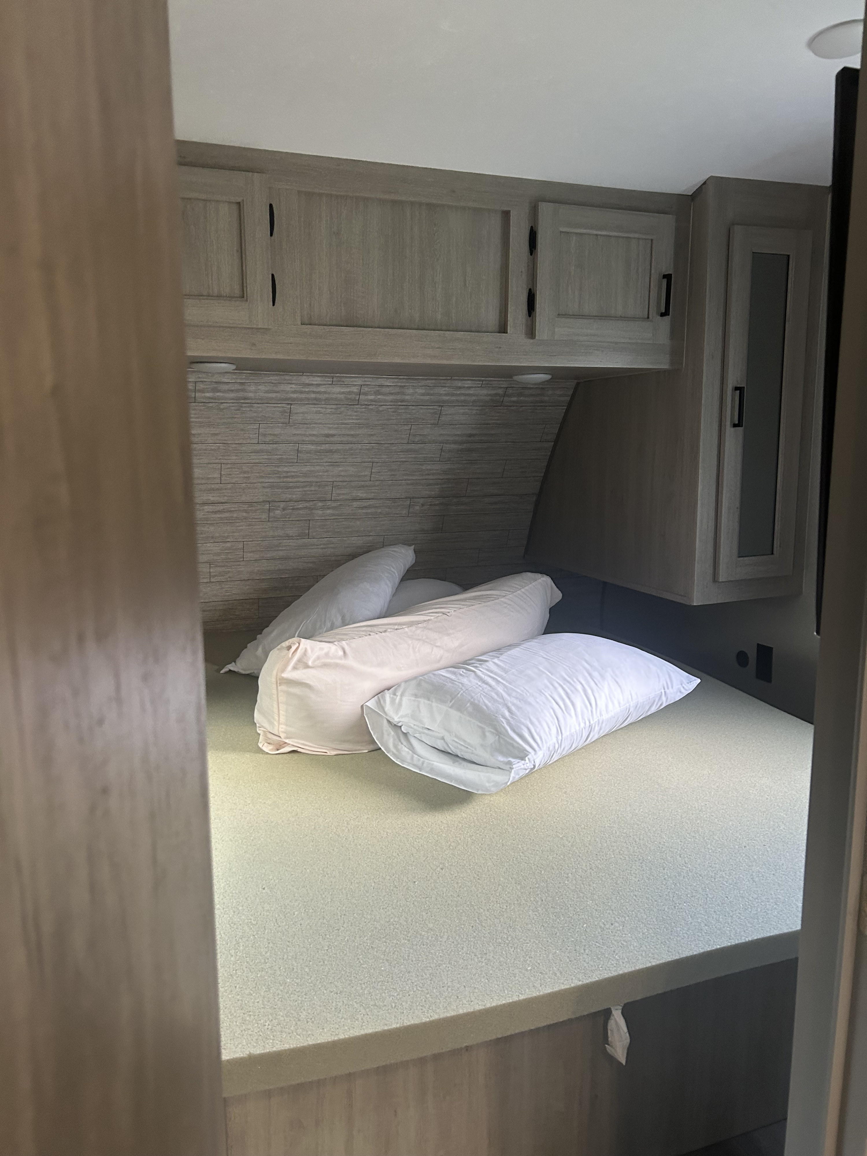 Cozy RV rental bedroom features a compact bed with pillows, a textured headboard, and wood-grain overhead cabinets.. Coachmen Catalina 2024