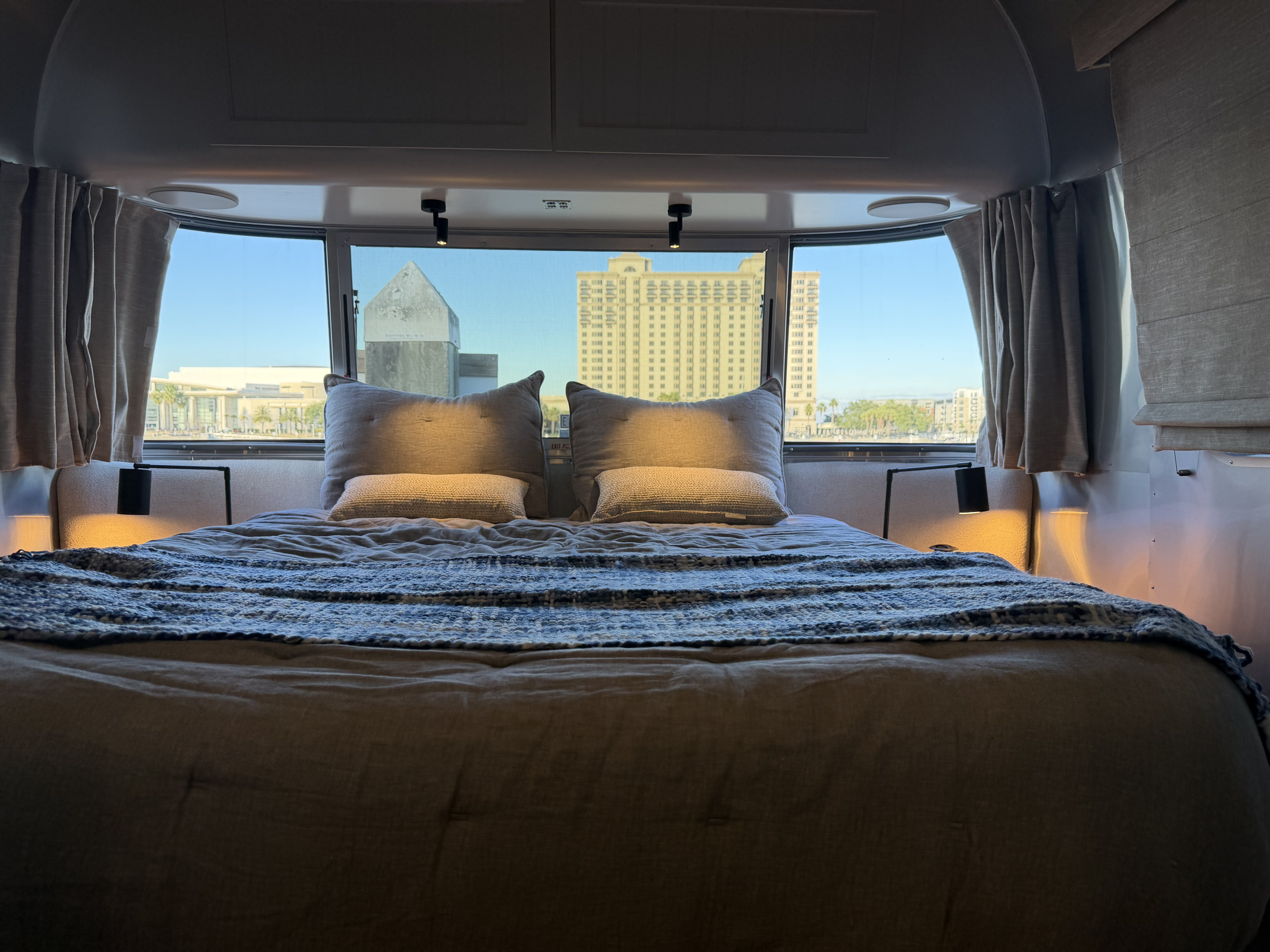 Cozy rv rental bedroom with plush bed, modern lighting, and a panoramic window view of a city waterfront, perfect for travelers.. Airstream Pottery Barn 2024