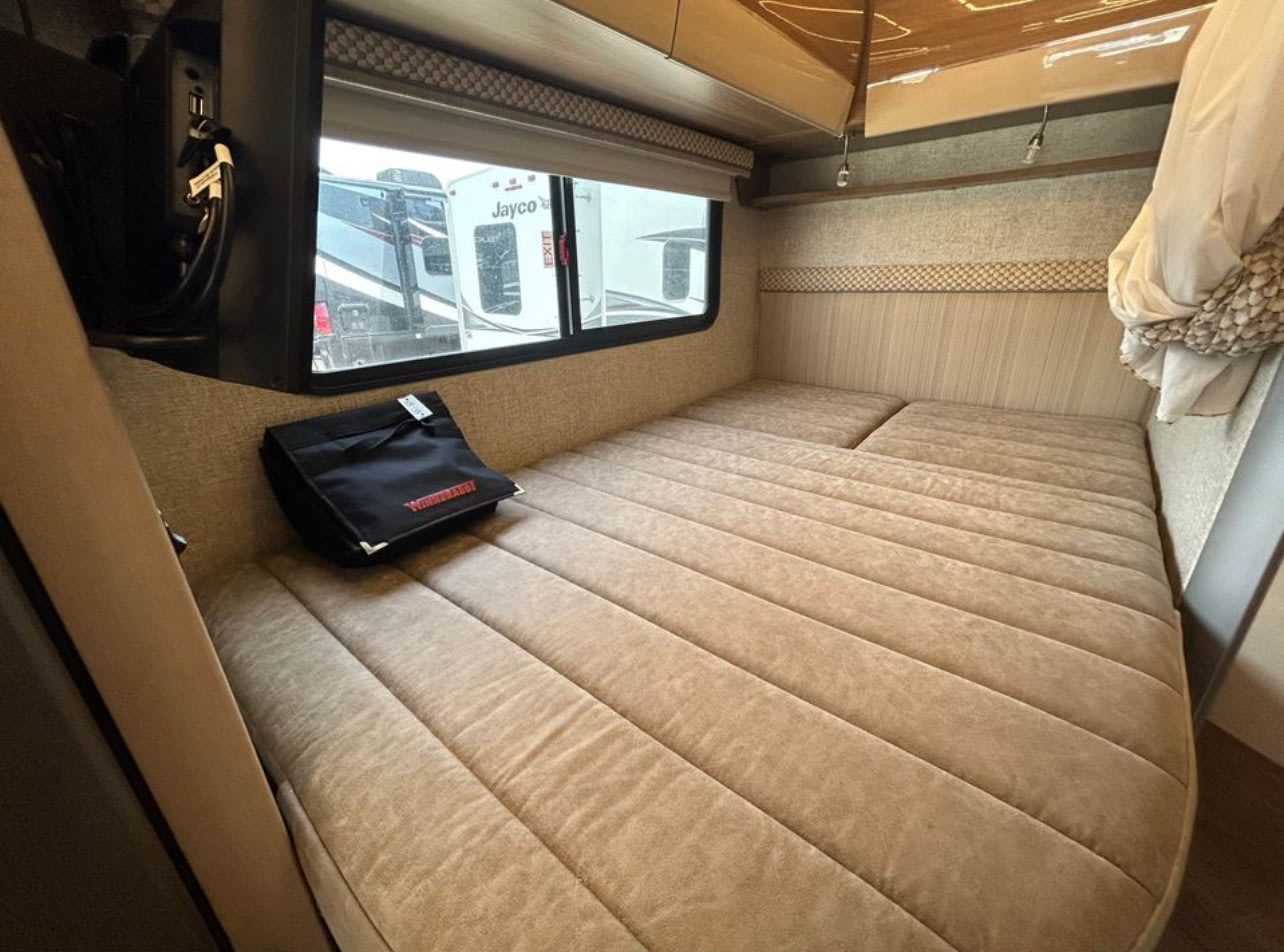 Inside an RV rental: a cozy fabric-cushioned bed with a window view of another RV, plus handy USB outlets.. Winnebago View 2018