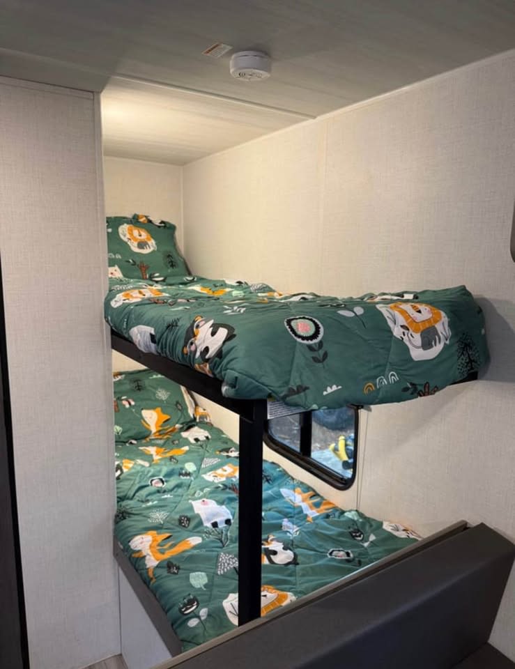 This RV rental features two cozy bunk beds with vibrant green animal-themed bedding, ideal for family trips. A smoke detector is visible.. Keystone RV Hideout Sport 2024