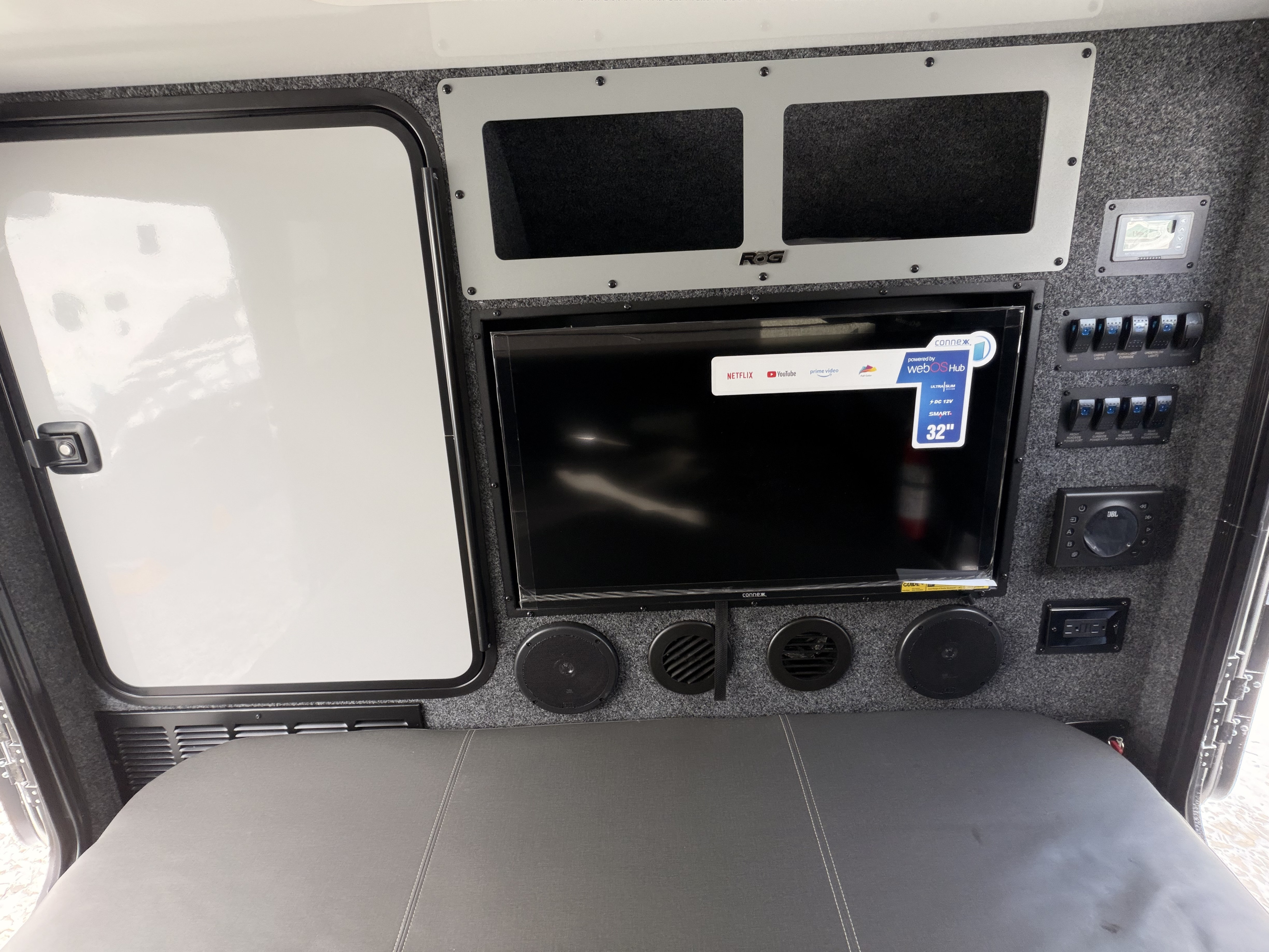 RV rental interior with a 32" Smart TV, JBL audio system, and a control panel for switches, above a gray bed area.. Encore ROG 12BH 2025