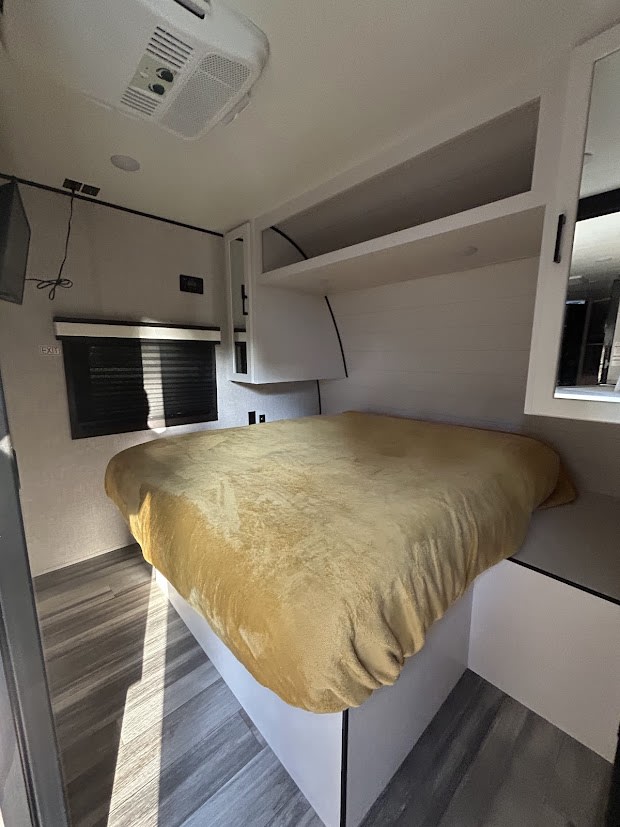 Cozy RV rental bedroom featuring a comfy bed with a golden blanket, overhead storage, AC unit, and wood-look flooring.. Jayco Jay Flight SLX 2025