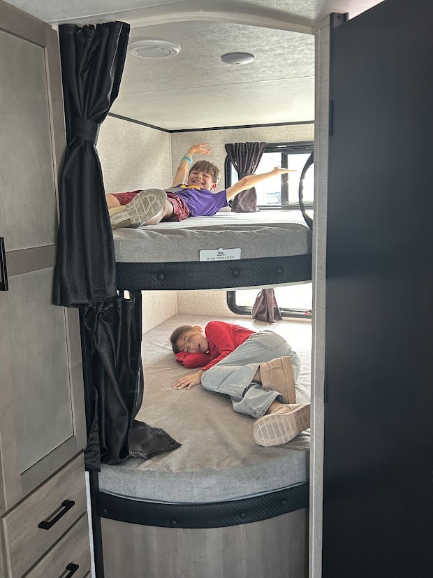 In an RV rental, two kids on bunk beds. One smiles and stretches on top, the other rests peacefully on the bottom.. Jayco Jay Flight SLX 2025