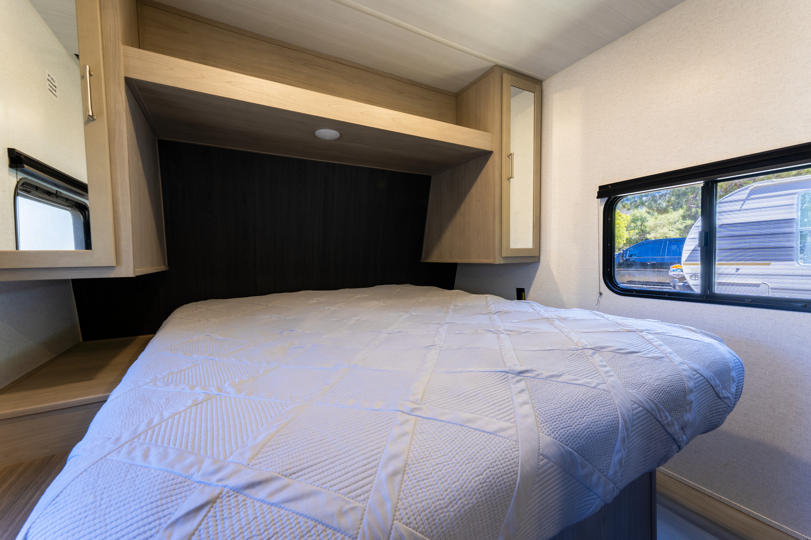 This RV rental bedroom features a comfy white bed, overhead cabinets, and a window looking out, ideal for travel trailer trips.. Keystone Springdale 2024