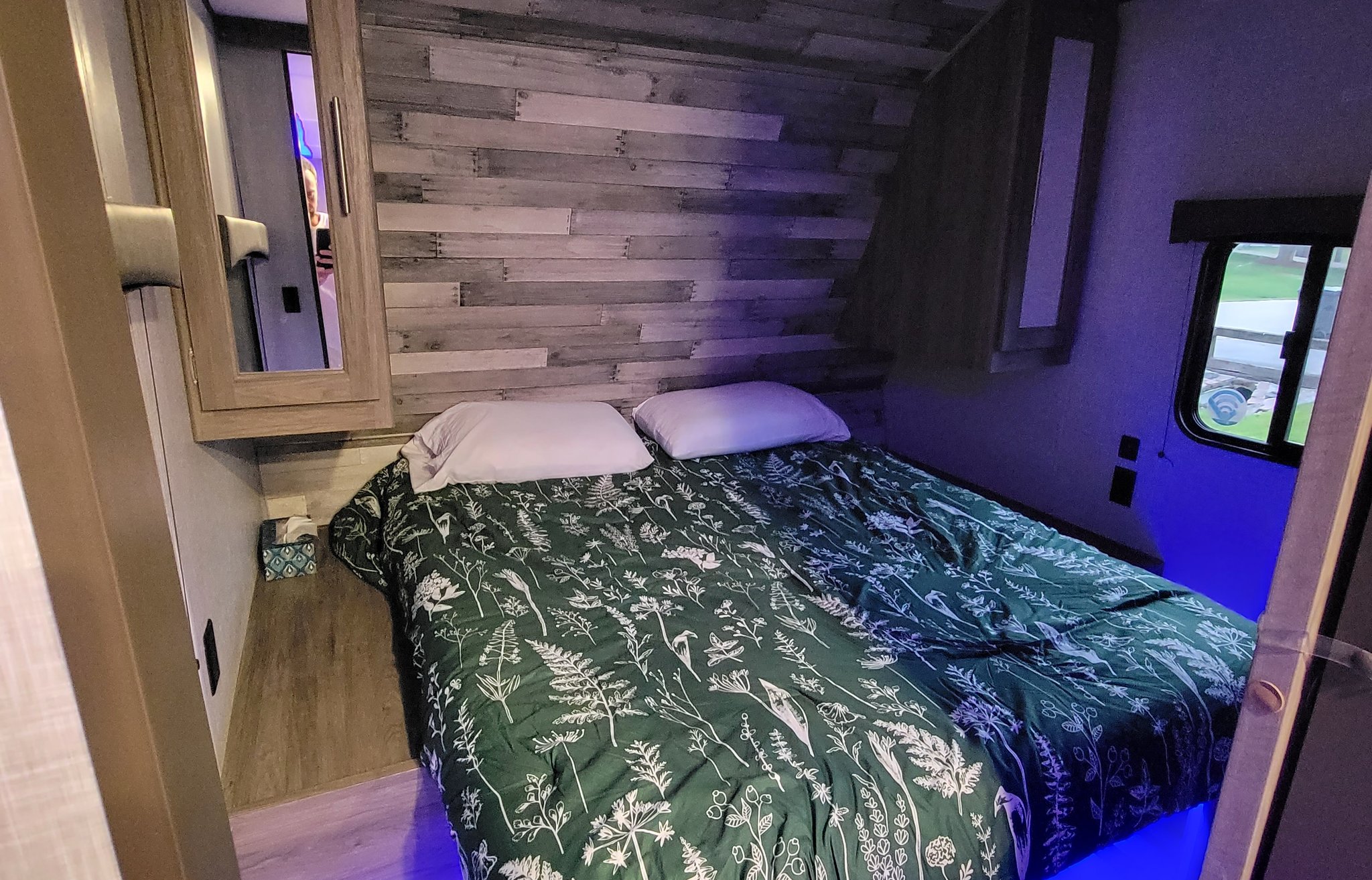 This cozy RV rental bedroom features a bed with a green floral comforter, rustic wood wall, and ambient blue LED lighting.. Crossroads RV Zinger Lite 2022