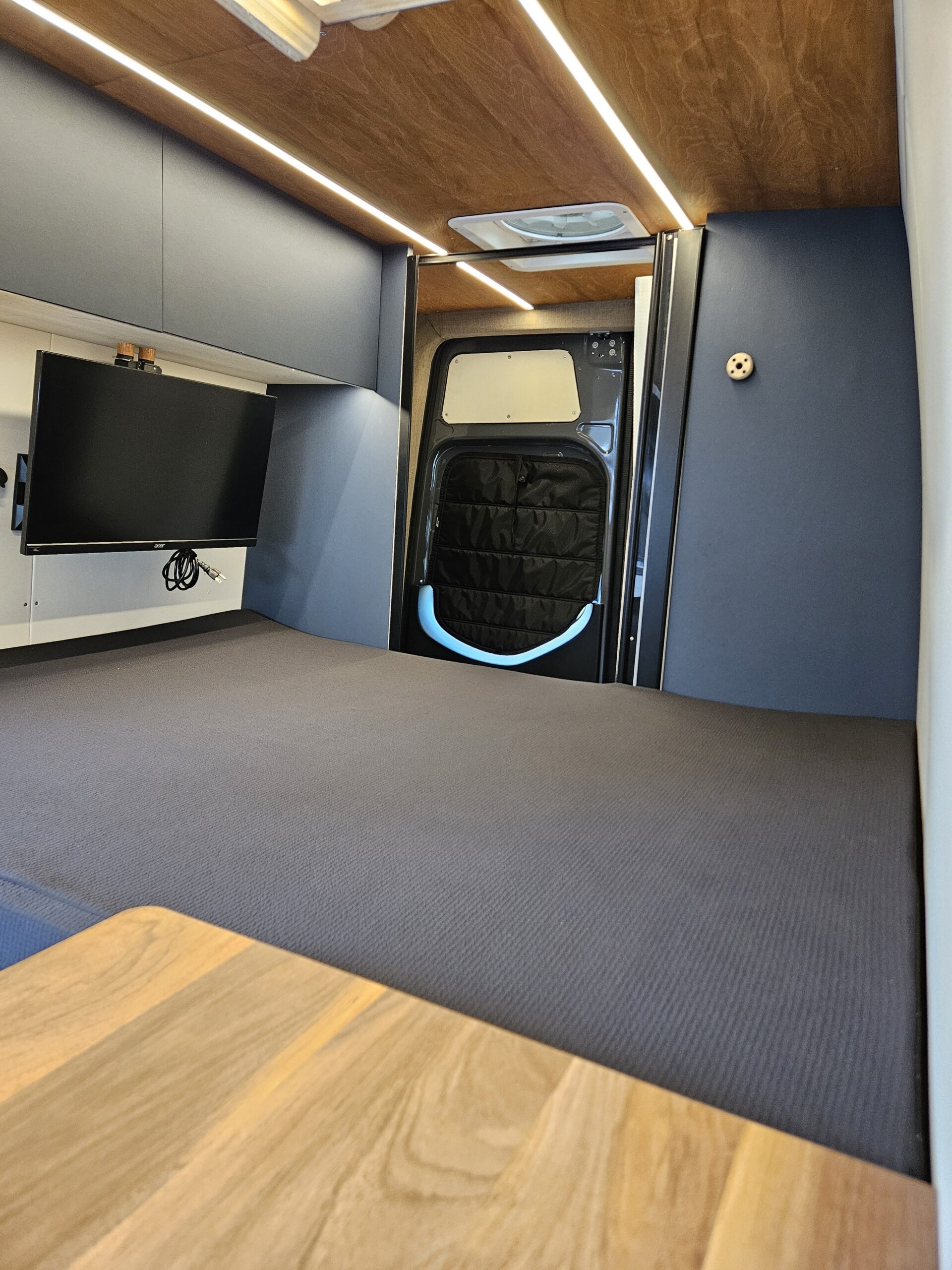 Inside a modern RV rental: comfy bed, wall-mounted TV, sleek dark cabinetry, and strip lighting on a wooden ceiling.. Venturhaus Sprinter 2022