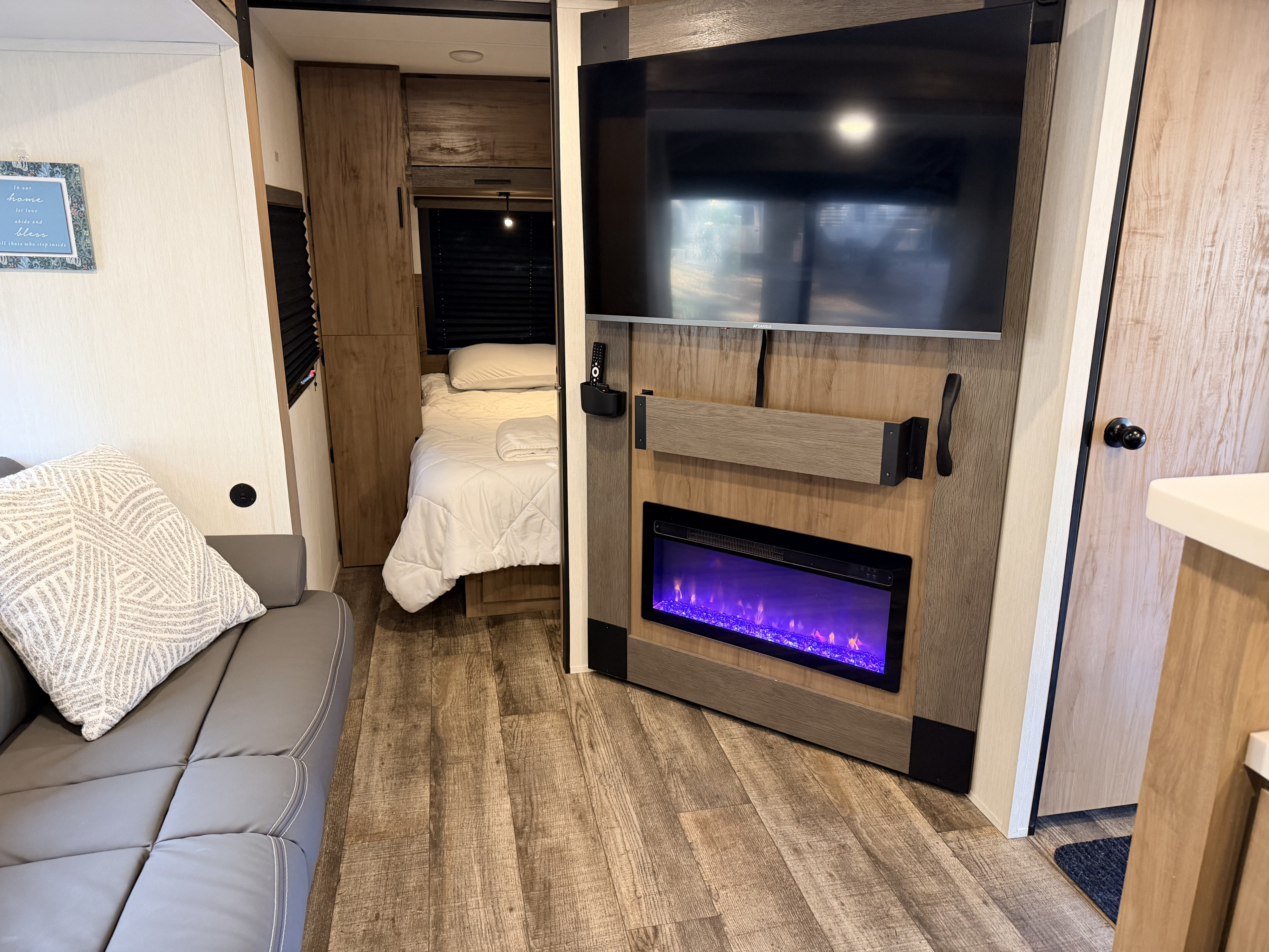 Cozy RV rental interior featuring a gray sofa, wood floors, a wall-mounted TV above an electric fireplace, and a bed.. Forest River Campsite Reserve 2025