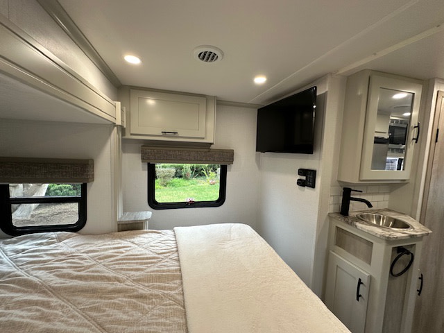 Experience comfort in this RV rental bedroom. It features a bed, two windows, a mounted TV, and a convenient sink vanity.. DynaMax iSata3 2025